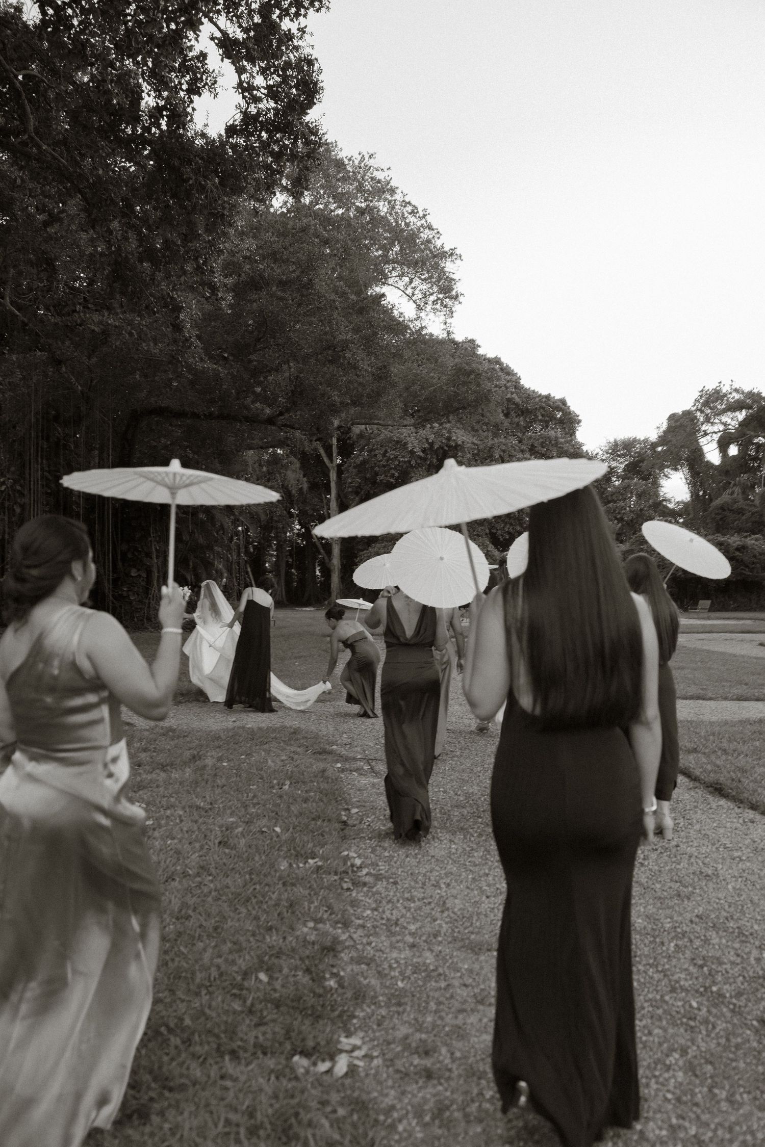 Black and white photo of women in elegant dresses carrying white parasols walking through a lush garden path.