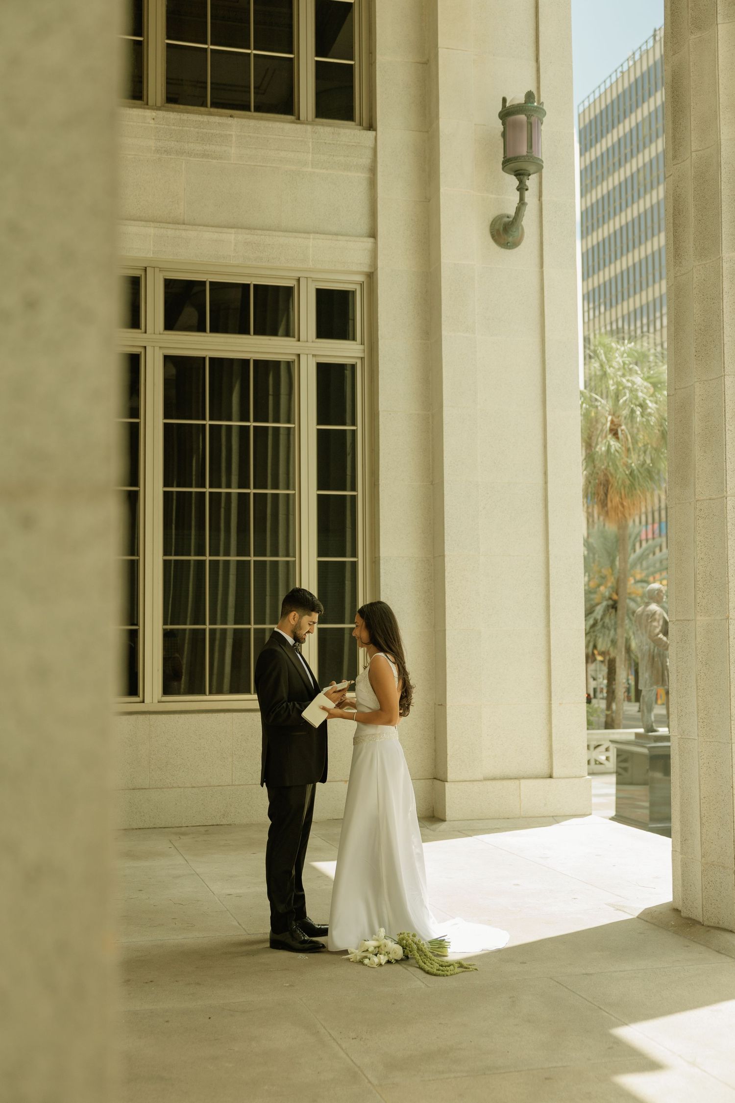 A bride and groom exchange vows between grand columns of a classical building, with palm trees visible in the background.