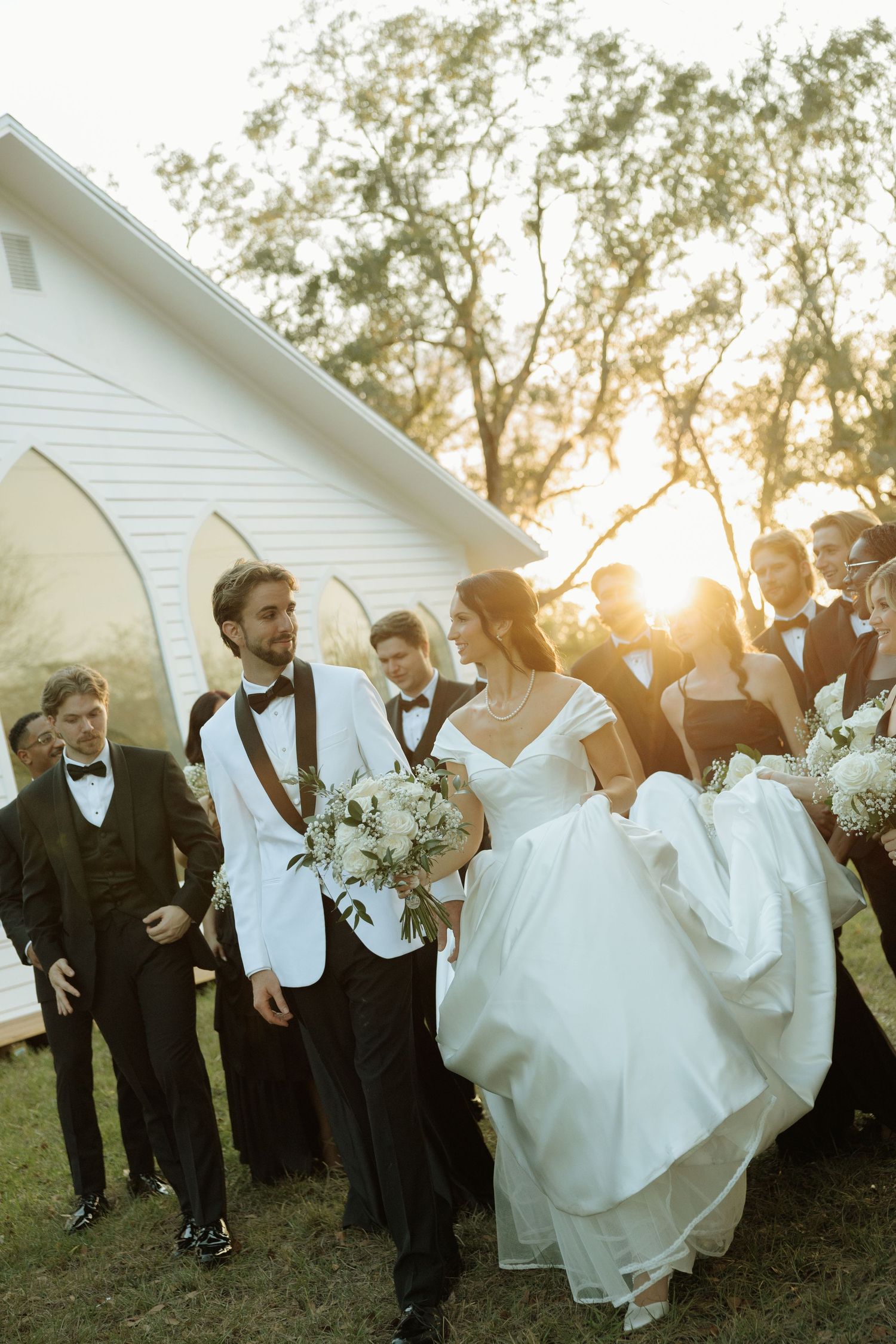 Bride and groom walk with wedding party outside white chapel at golden hour sunset.