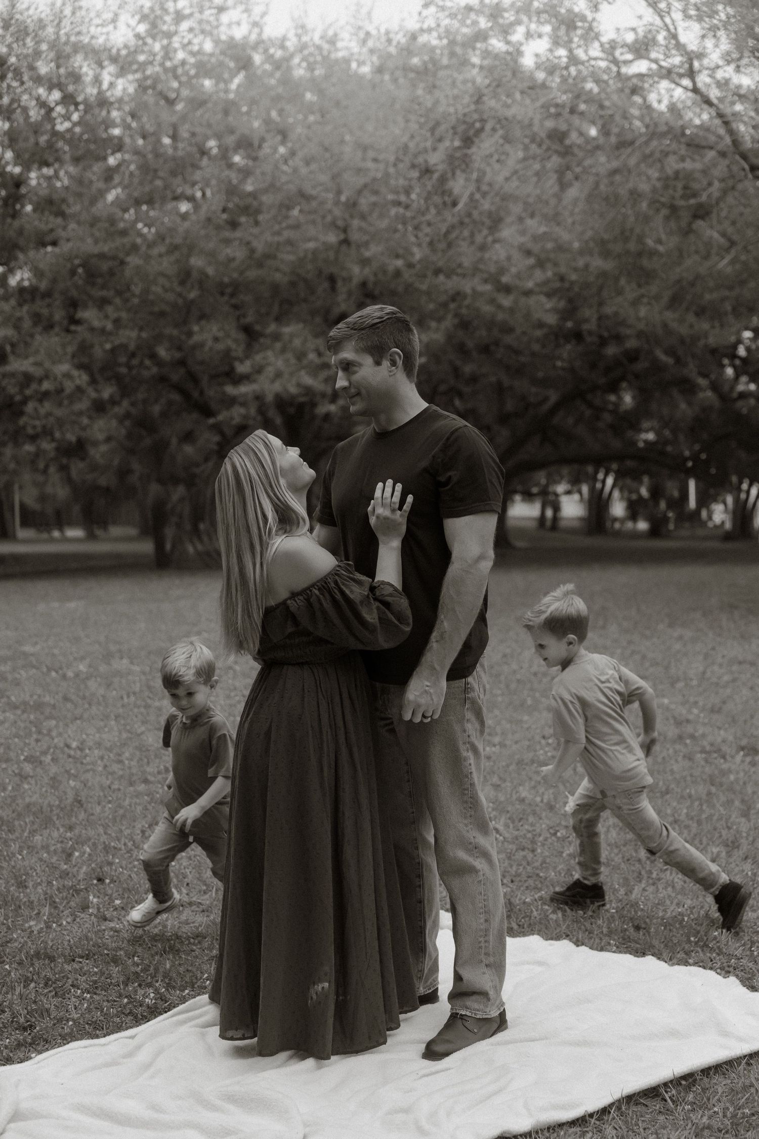Black and white photo series showing a toddler playing and walking on a blanket outdoors while adults watch nearby.