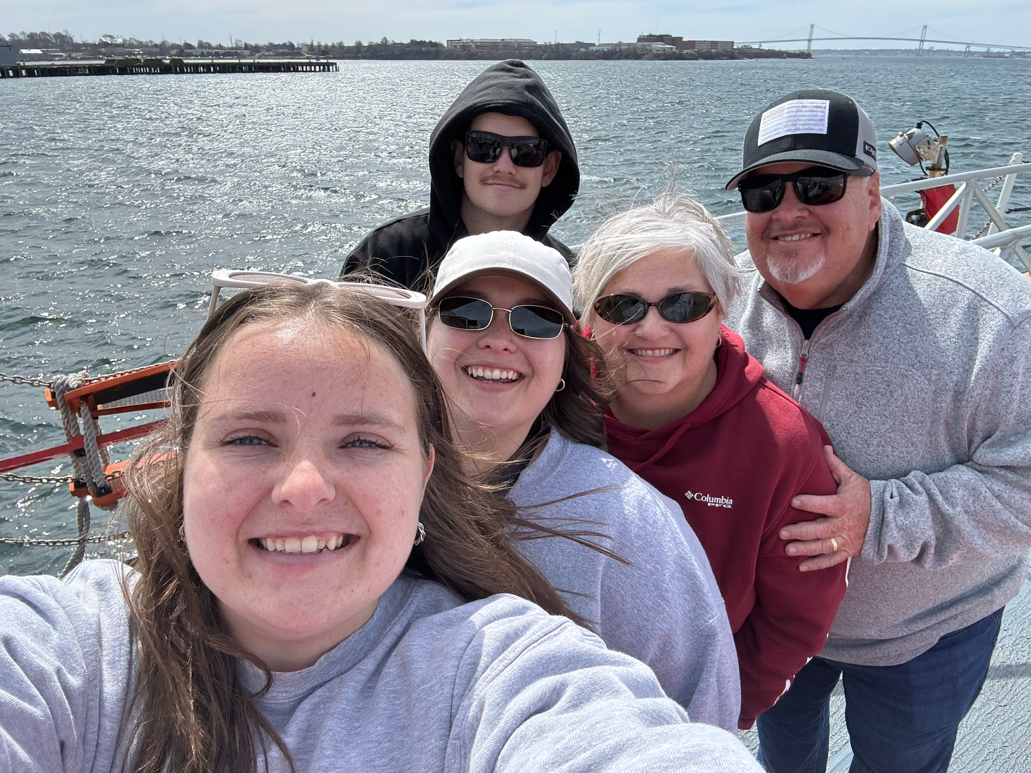 A group takes a selfie together while on a boat with the ocean and shoreline visible in the background.