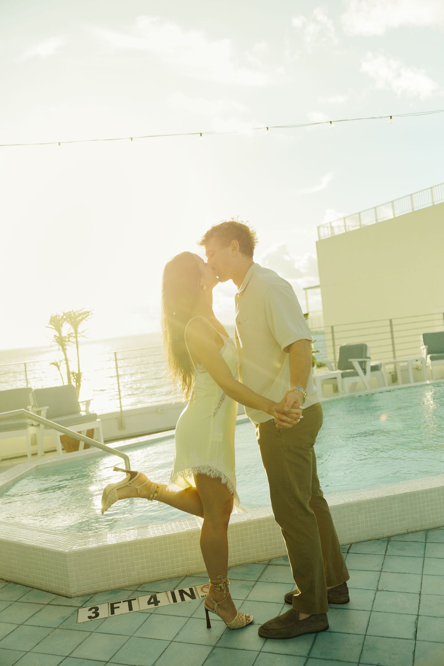 Couple kissing romantically by a rooftop pool at sunset with ocean views in the background.