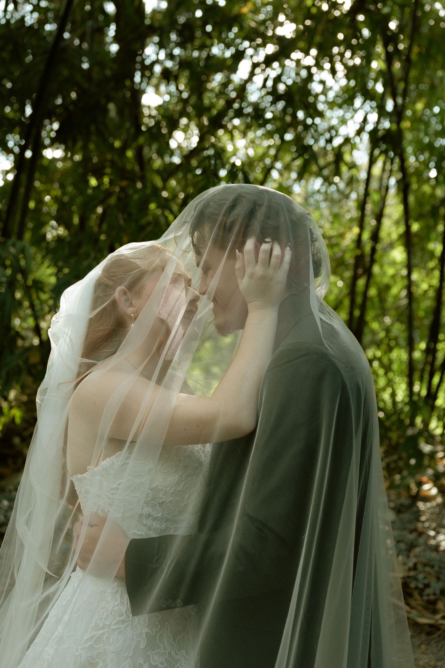 Bride and groom share an intimate moment under a flowing white veil surrounded by lush green trees.