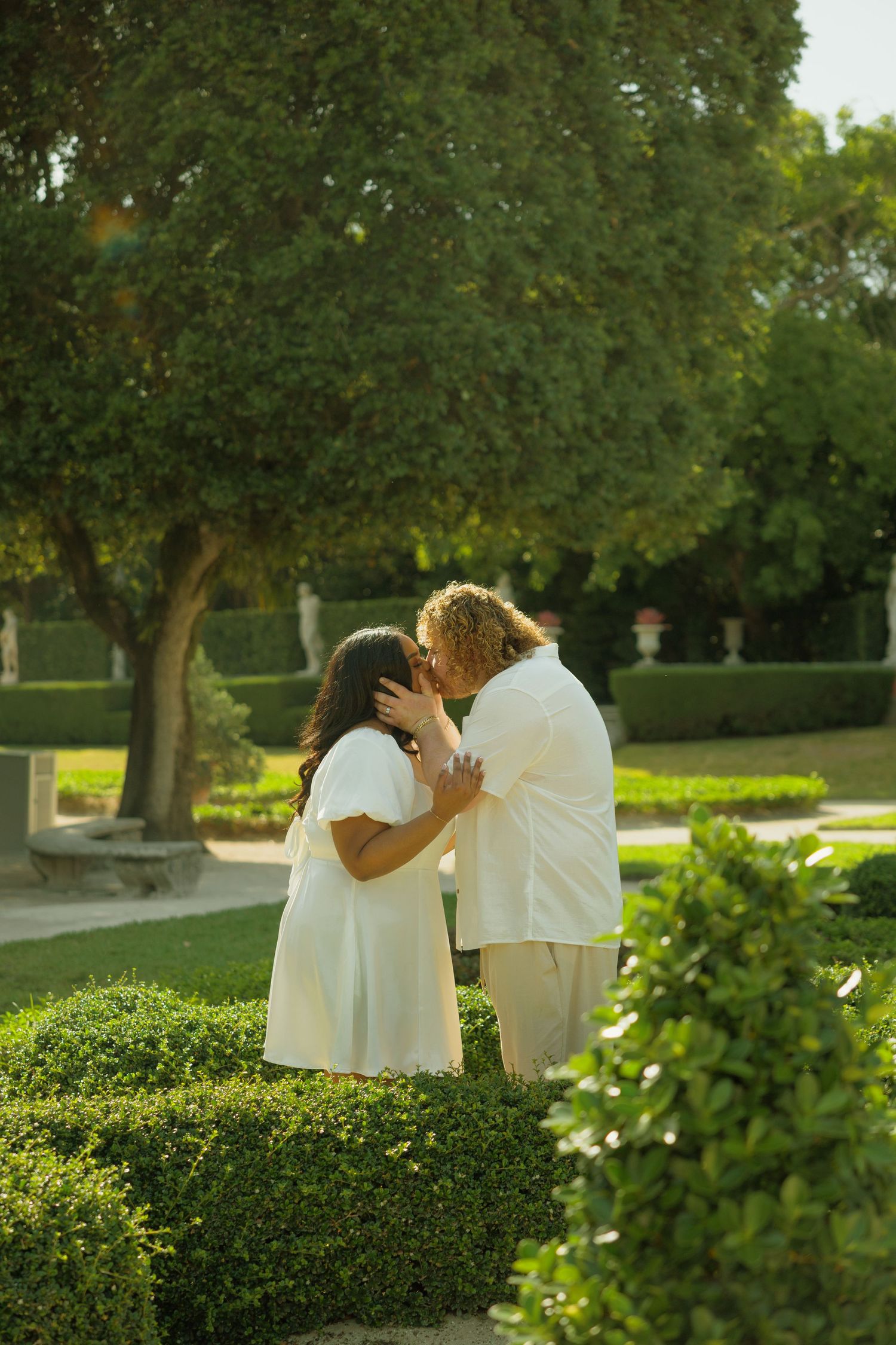 Couple in white outfits sharing a romantic kiss in a lush green garden with manicured hedges and trees.