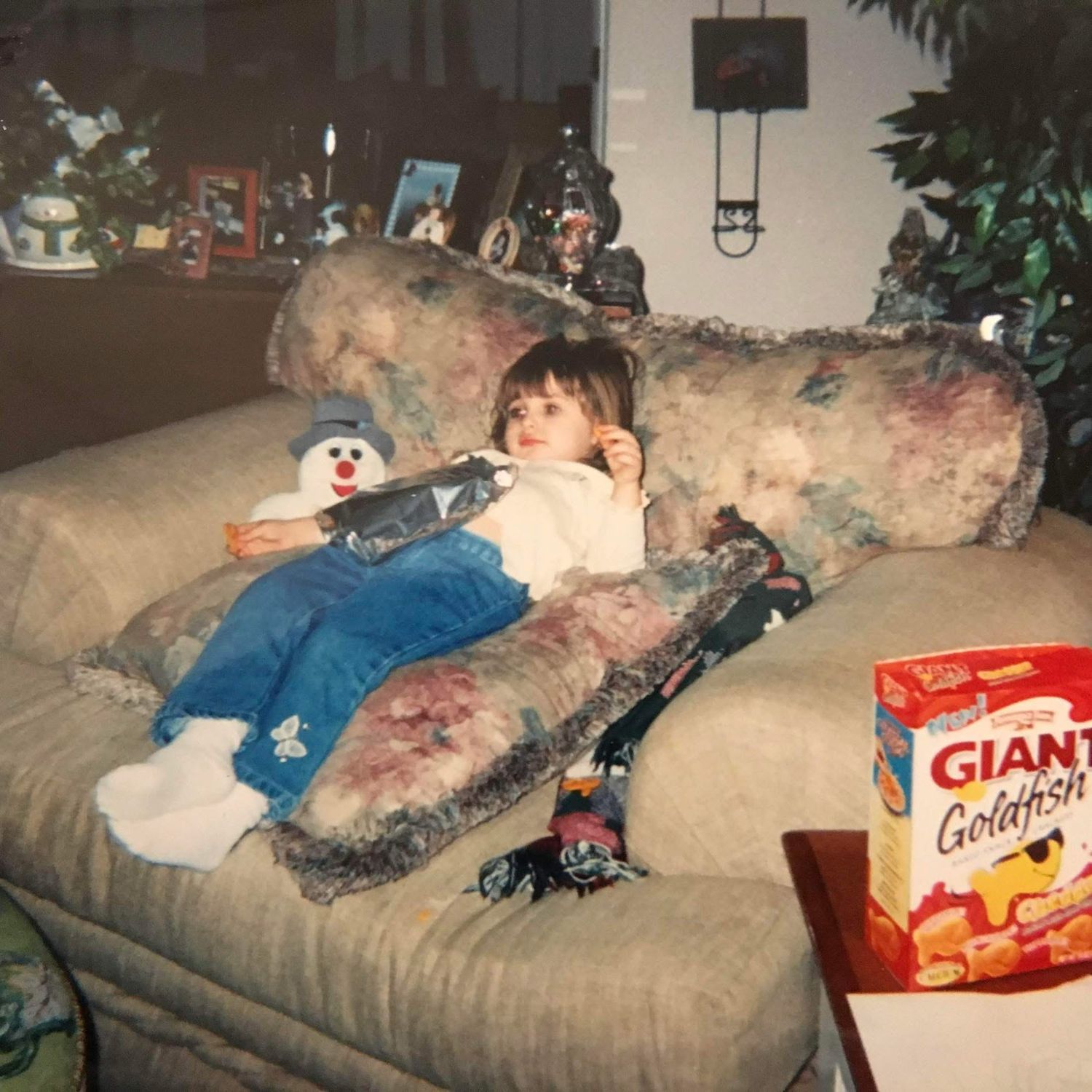Person lounging on gray couch with snacks and entertainment visible in a dimly lit room setting.