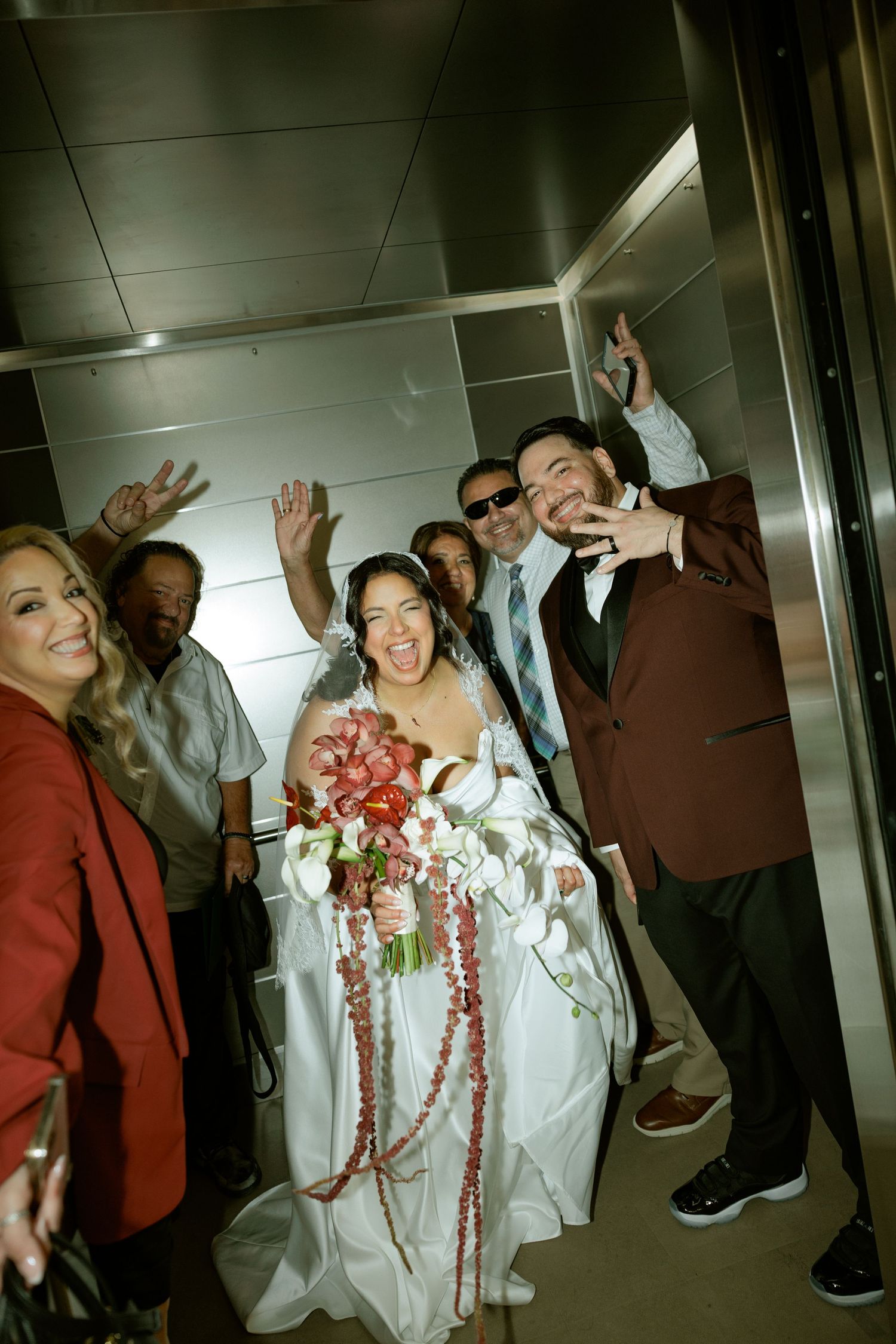 Bride in white gown holding floral bouquet celebrates with groom and guests inside elevator at wedding reception.