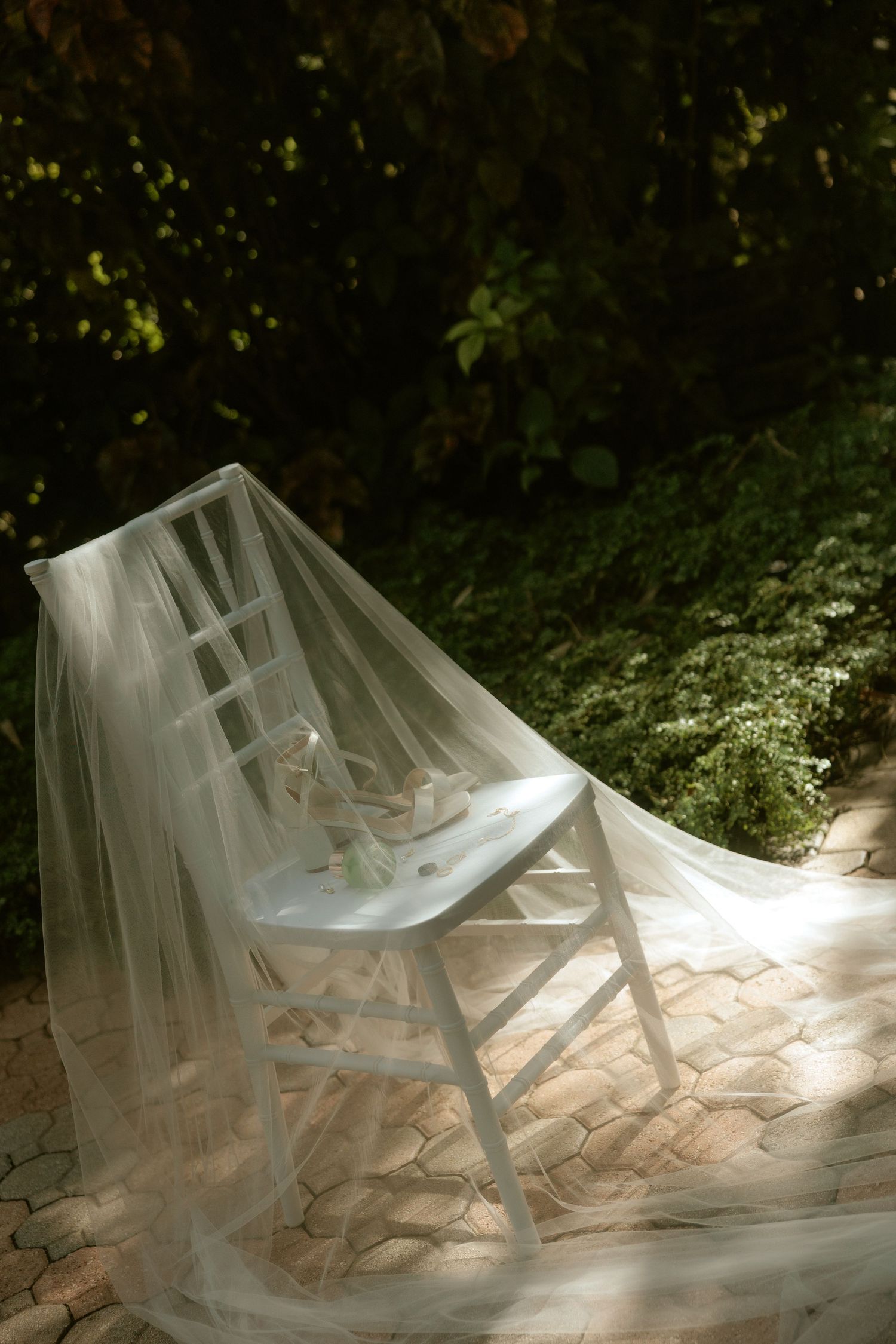 Elegant white bridal veil draped over a white wooden chair outdoors, surrounded by lush green foliage in soft sunlight.