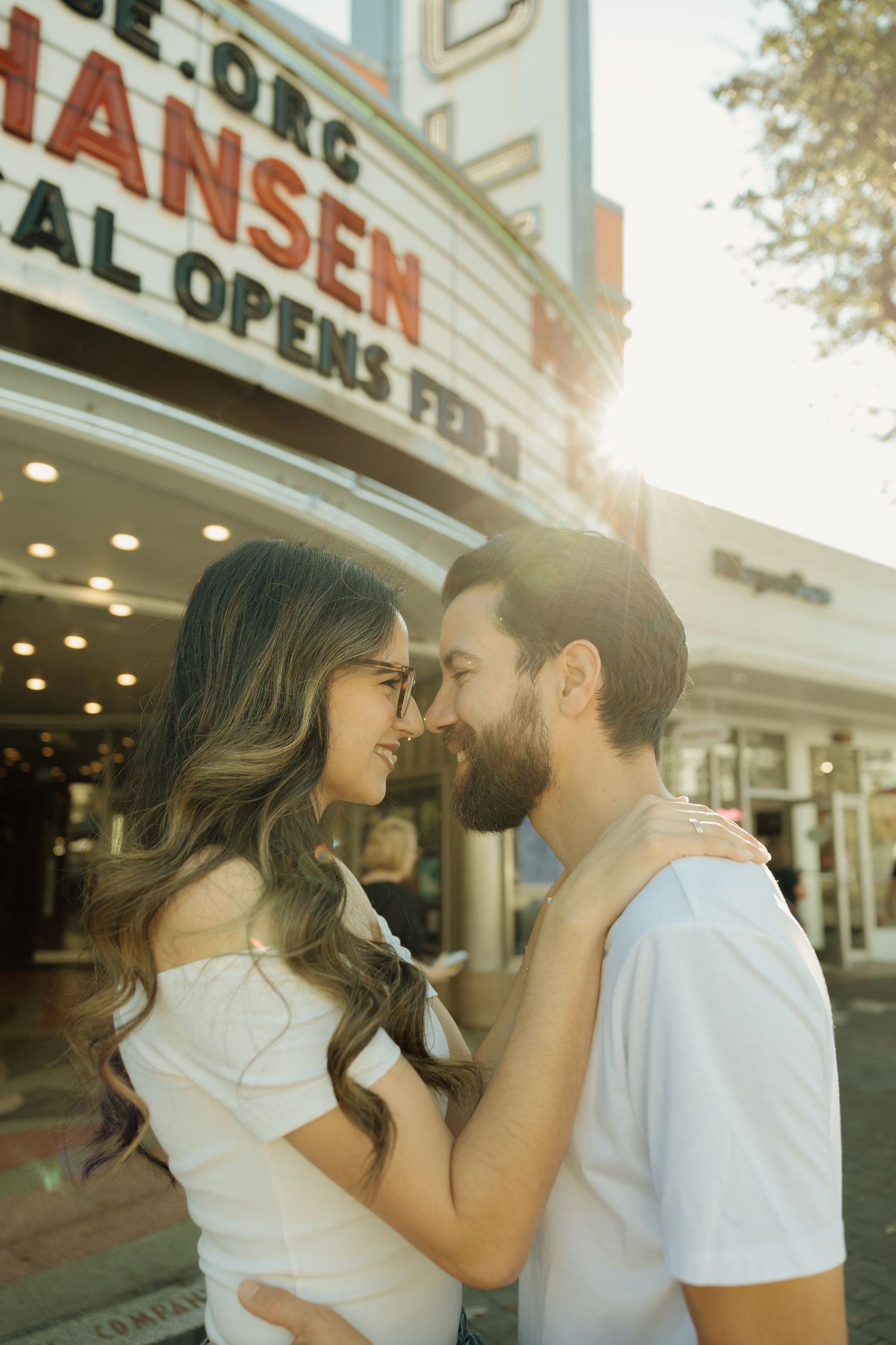 A couple shares a tender moment in front of a vintage movie theater marquee on a sunny afternoon.