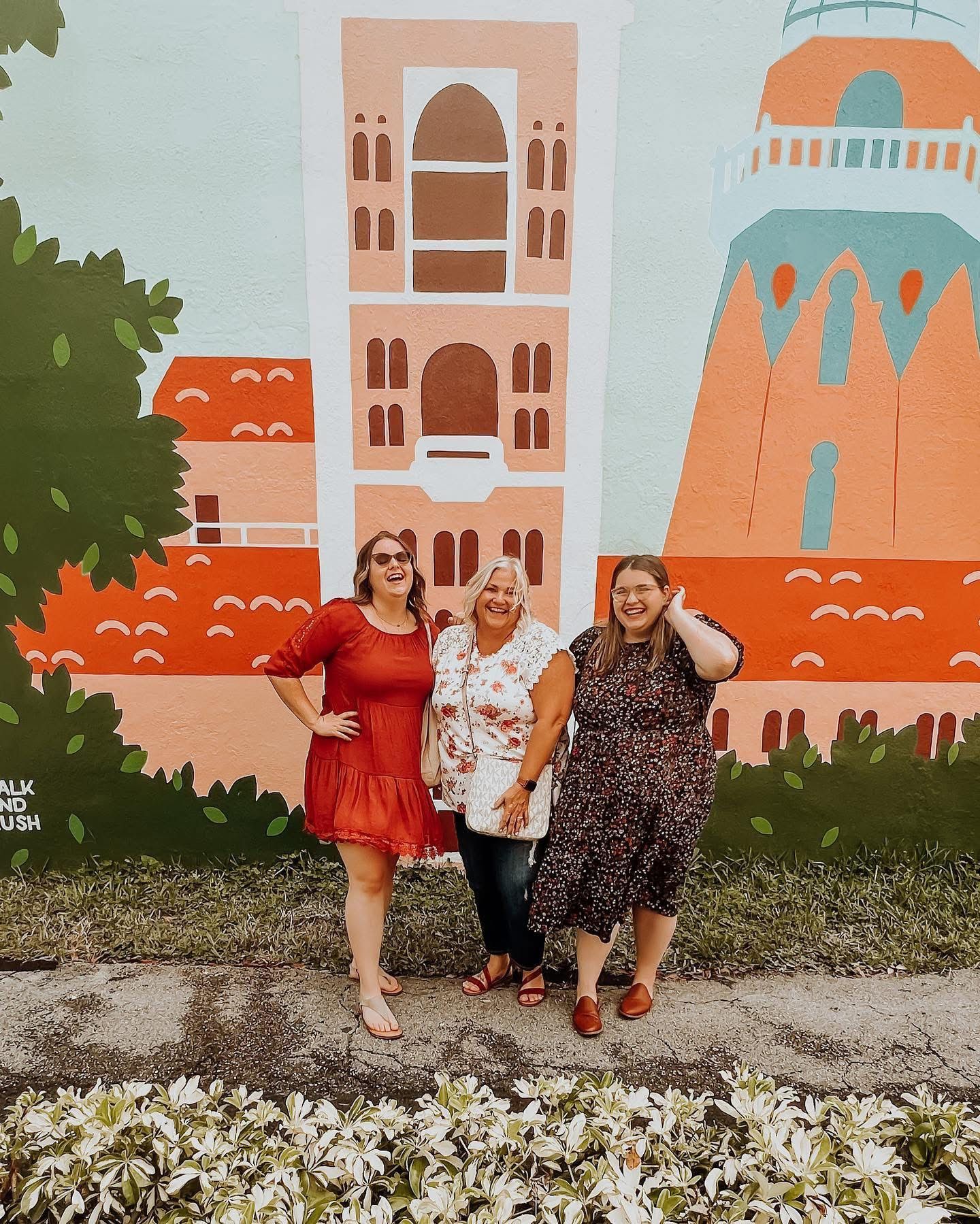 Three people pose in front of a colorful mural depicting orange and pink buildings with architectural details and foliage.