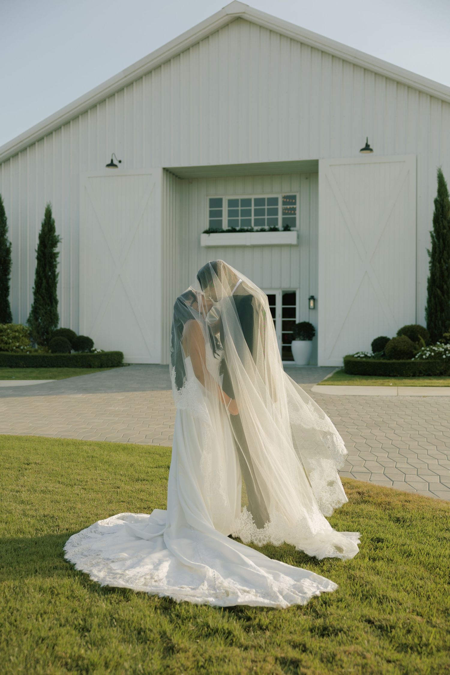Two brides share a kiss under a flowing veil in front of a white barn wedding venue on a green lawn.