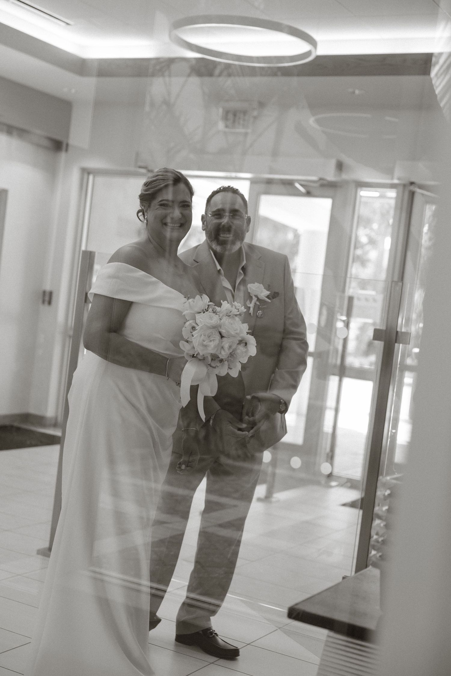 Black and white photo of smiling bride holding bouquet and groom in suit posing together in elegant venue lobby.