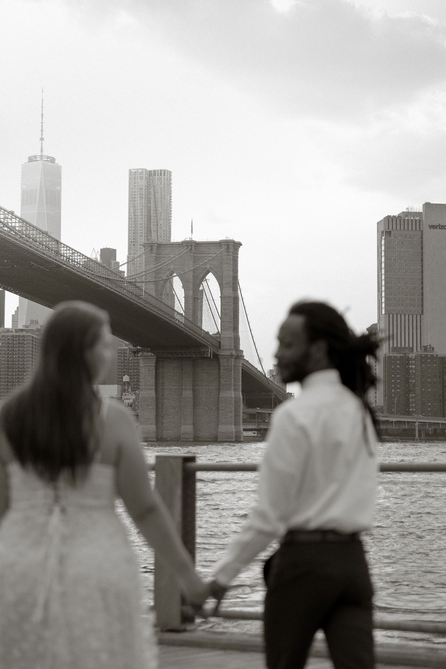 A couple stands silently by the water near the Brooklyn Bridge with the New York City skyline in the background.