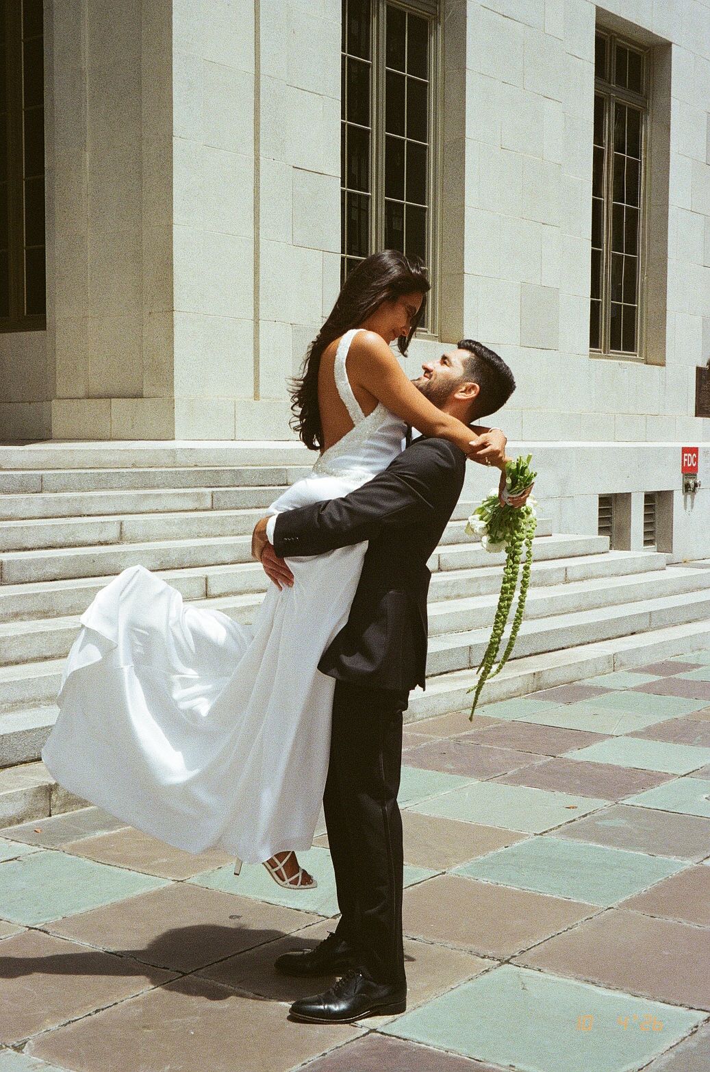Groom in black suit lifts bride in white gown for a kiss on steps of elegant building on wedding day.