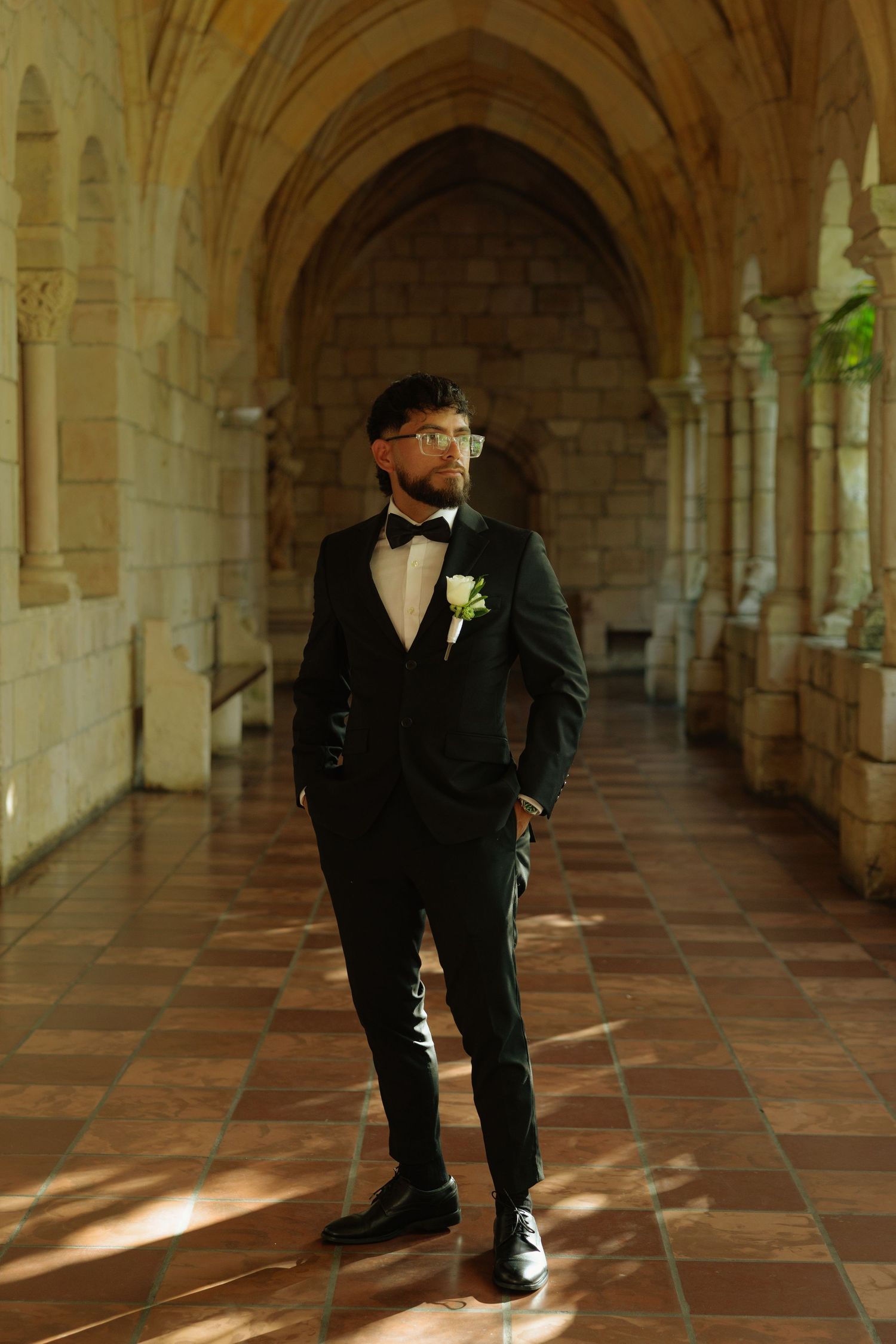 Dapper groom in black tuxedo with bow tie and boutonniere stands in elegant stone archway corridor.