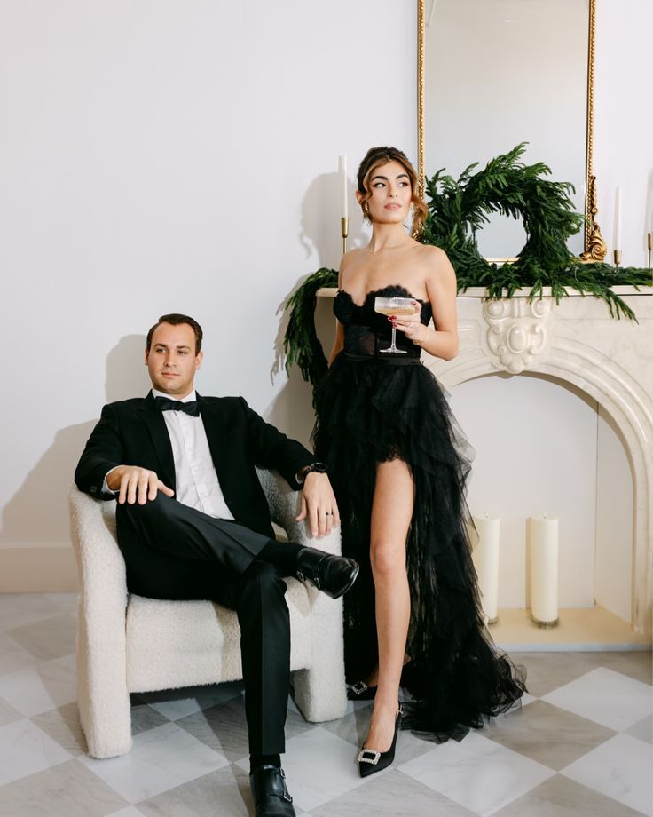 Elegant couple in black formal attire posing by a fireplace with greenery, man in tuxedo seated, woman in gown holding wine.