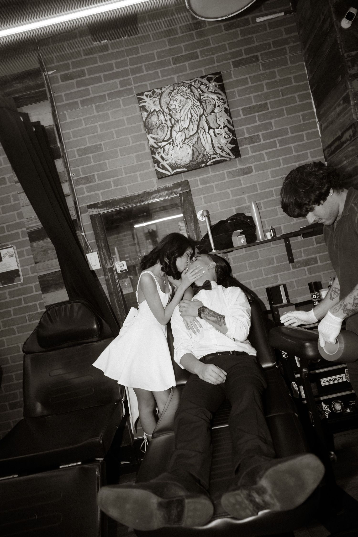 A black and white photograph shows people dancing and lounging in a brick-walled room with artwork hanging above.