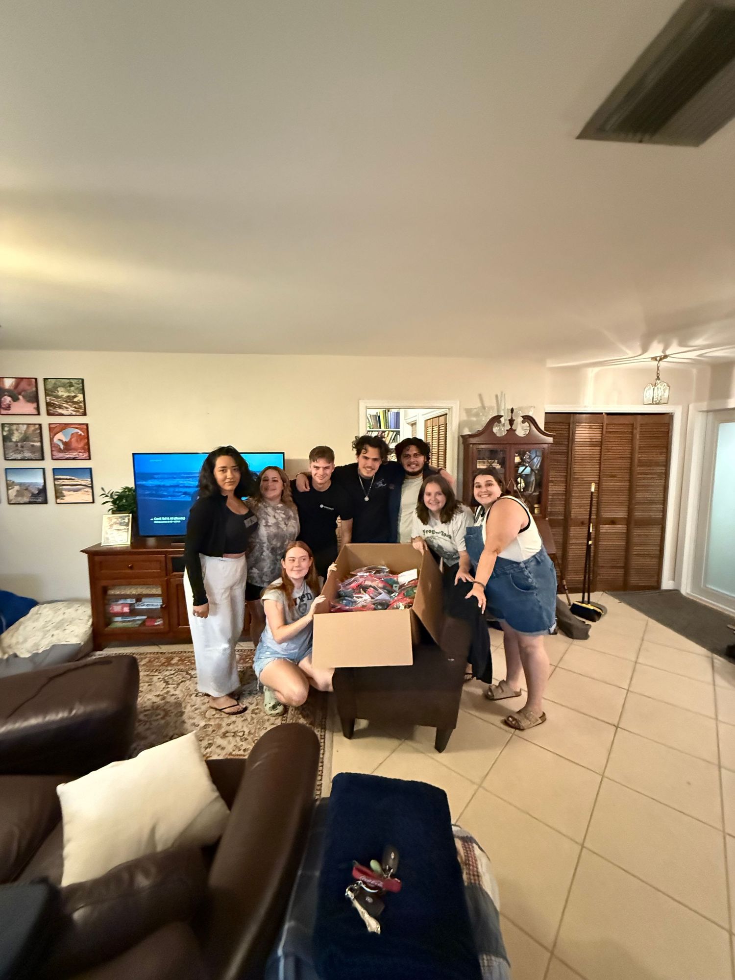 A group gathers around a coffee table in a living room with brown leather furniture and tiled floors.