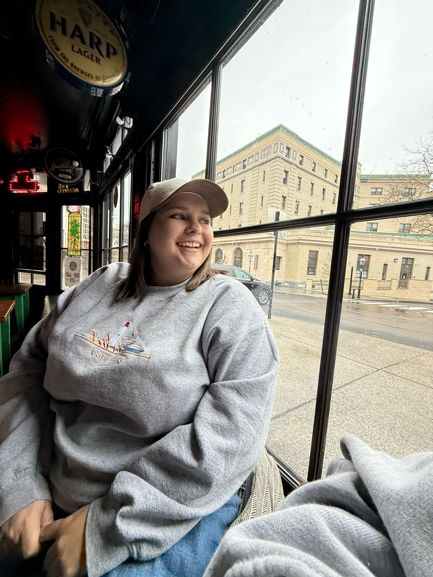 Someone wearing a gray sweatshirt and baseball cap sits by a window in a restaurant or cafe with city buildings visible outside.