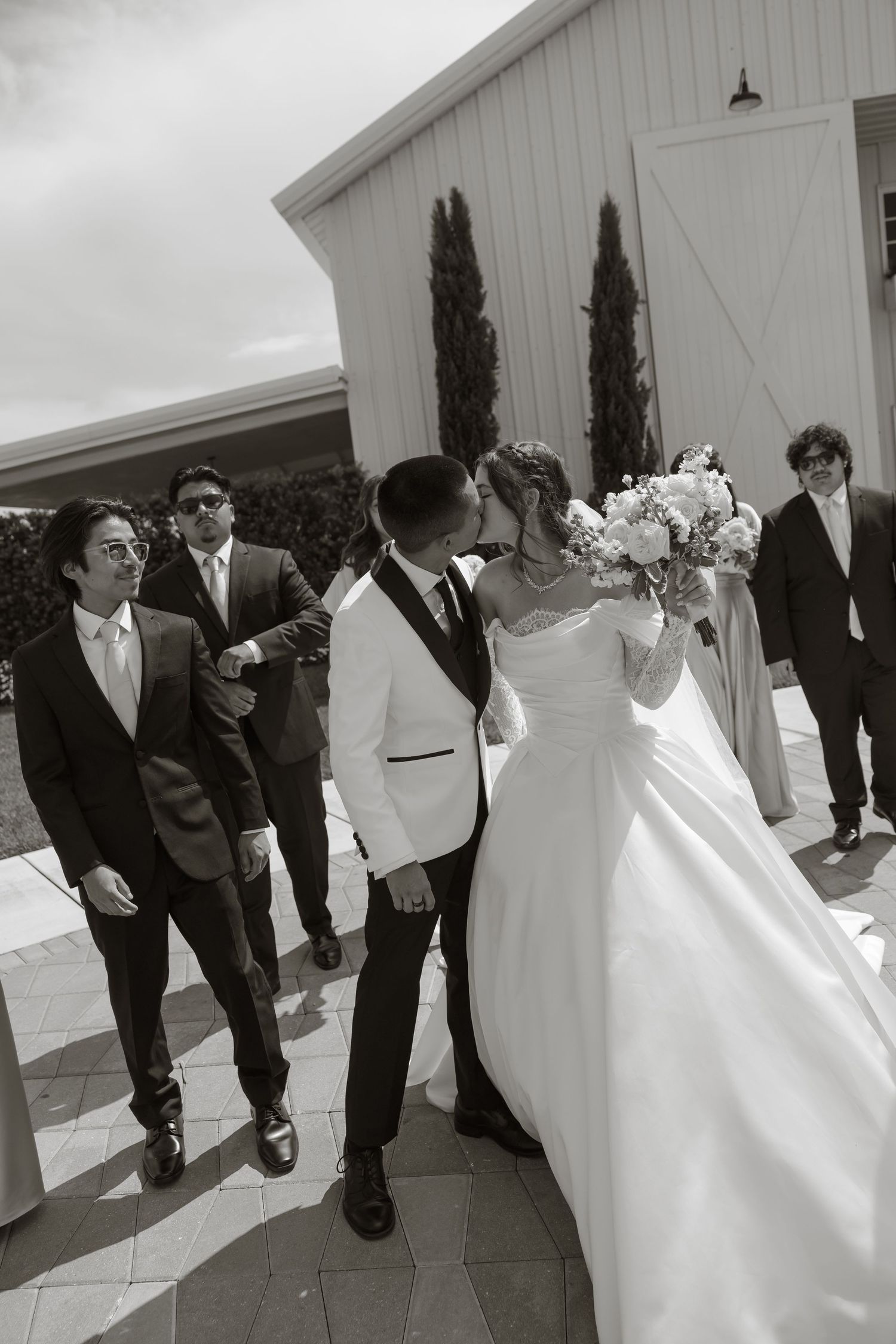 Black and white photo of a bride and groom kissing outside a white barn venue, surrounded by groomsmen and a bridesmaid.