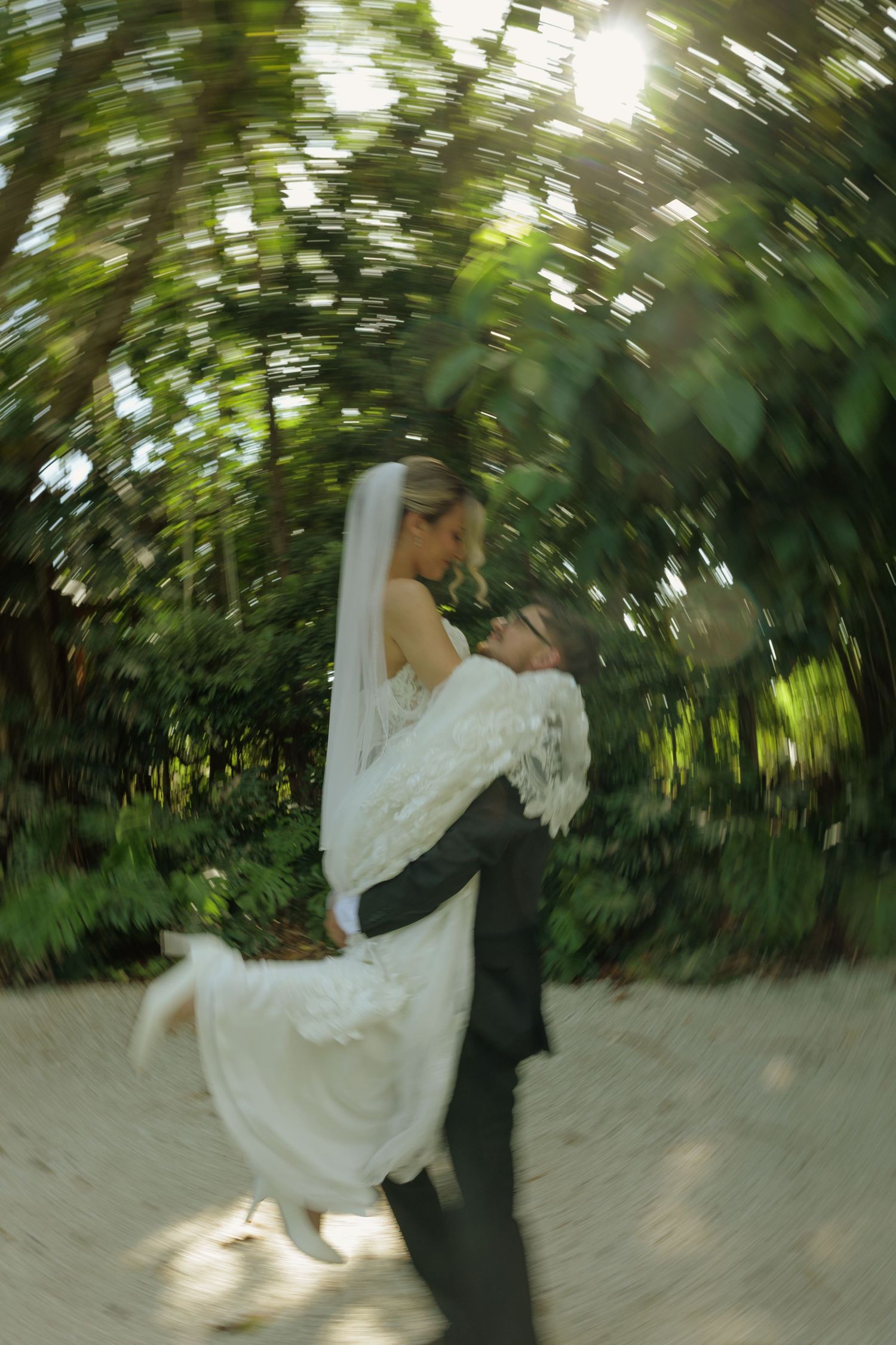 Groom lifting bride in white wedding dress and veil, spinning in lush green garden with dreamy motion blur effect.