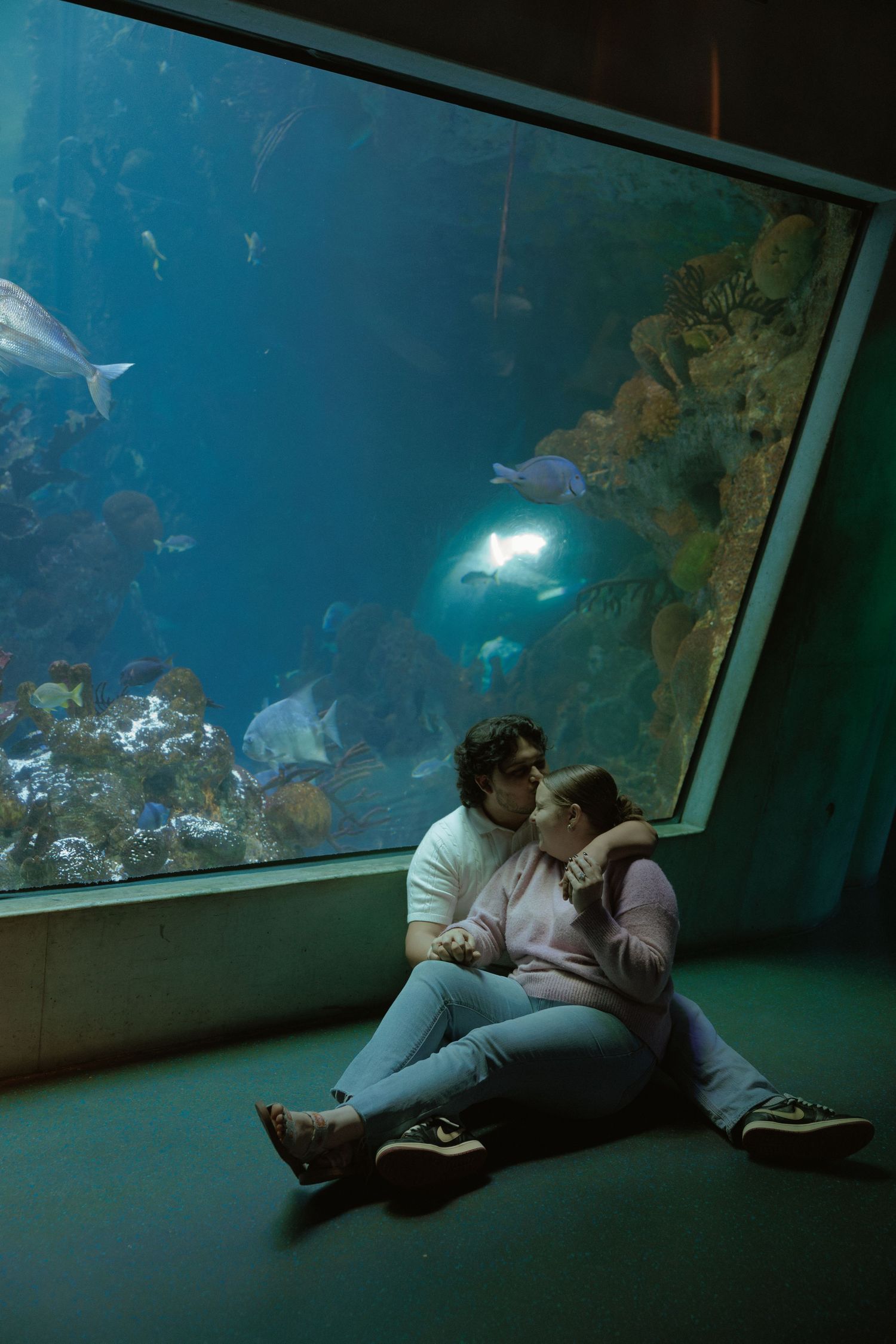 A couple shares a romantic moment while lying on the floor beneath an aquarium window with blue lighting.
