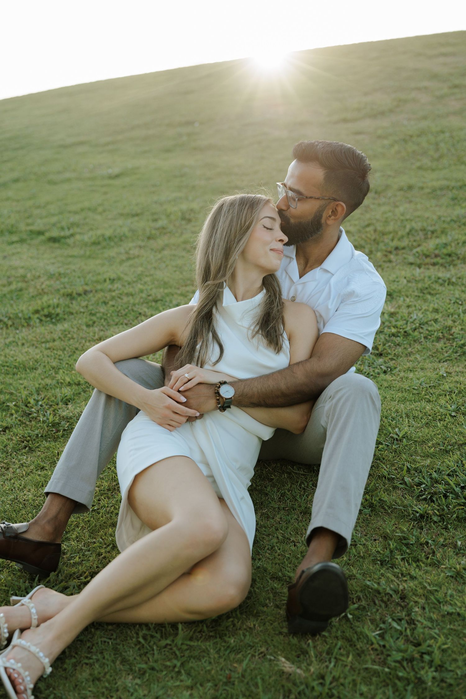 A couple shares an intimate moment while sitting together on grass during a sunny afternoon photoshoot in white attire.