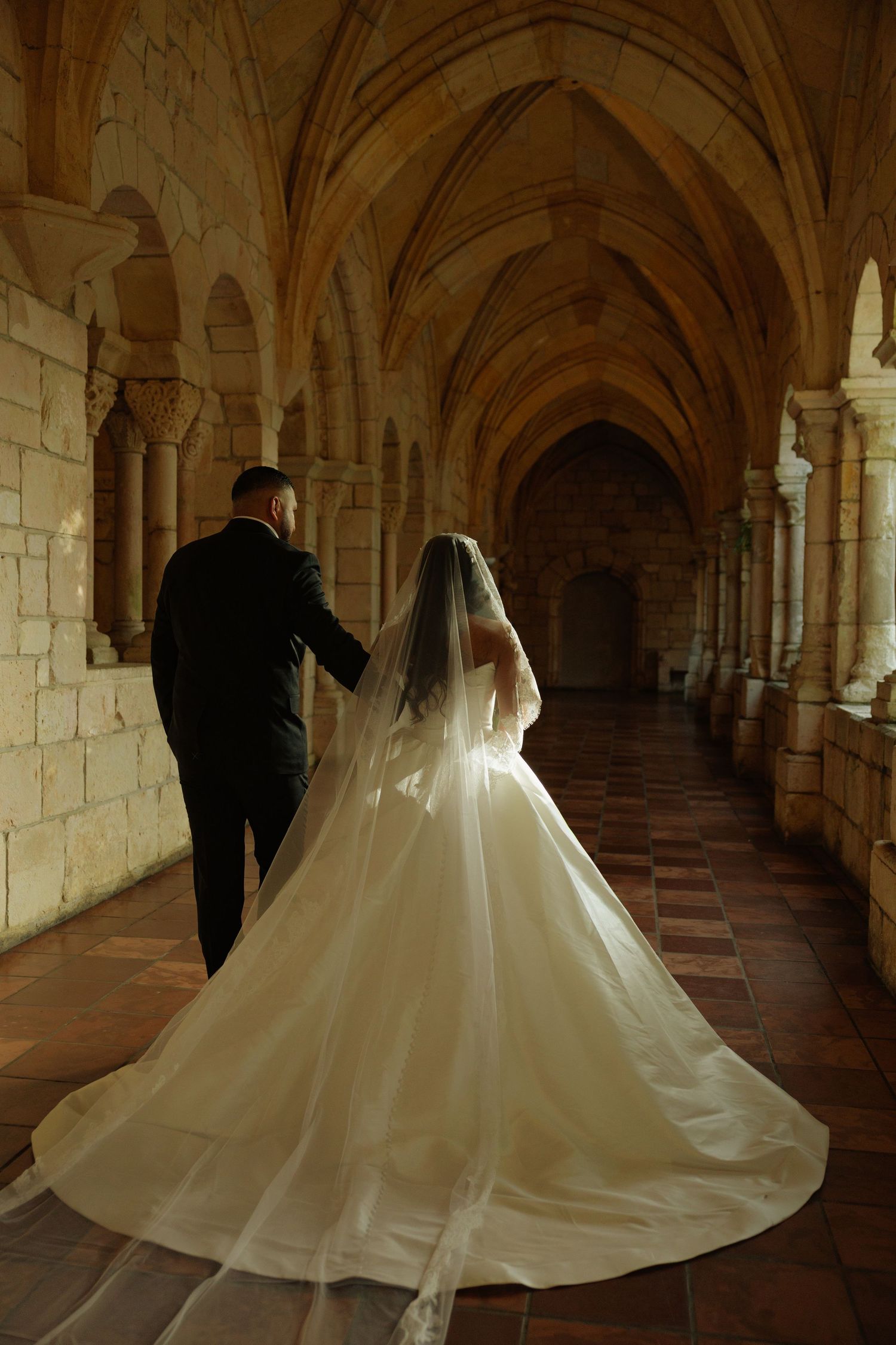 Bride and groom walking through a grand Gothic stone archway corridor, bride wearing flowing white gown with long veil and train.