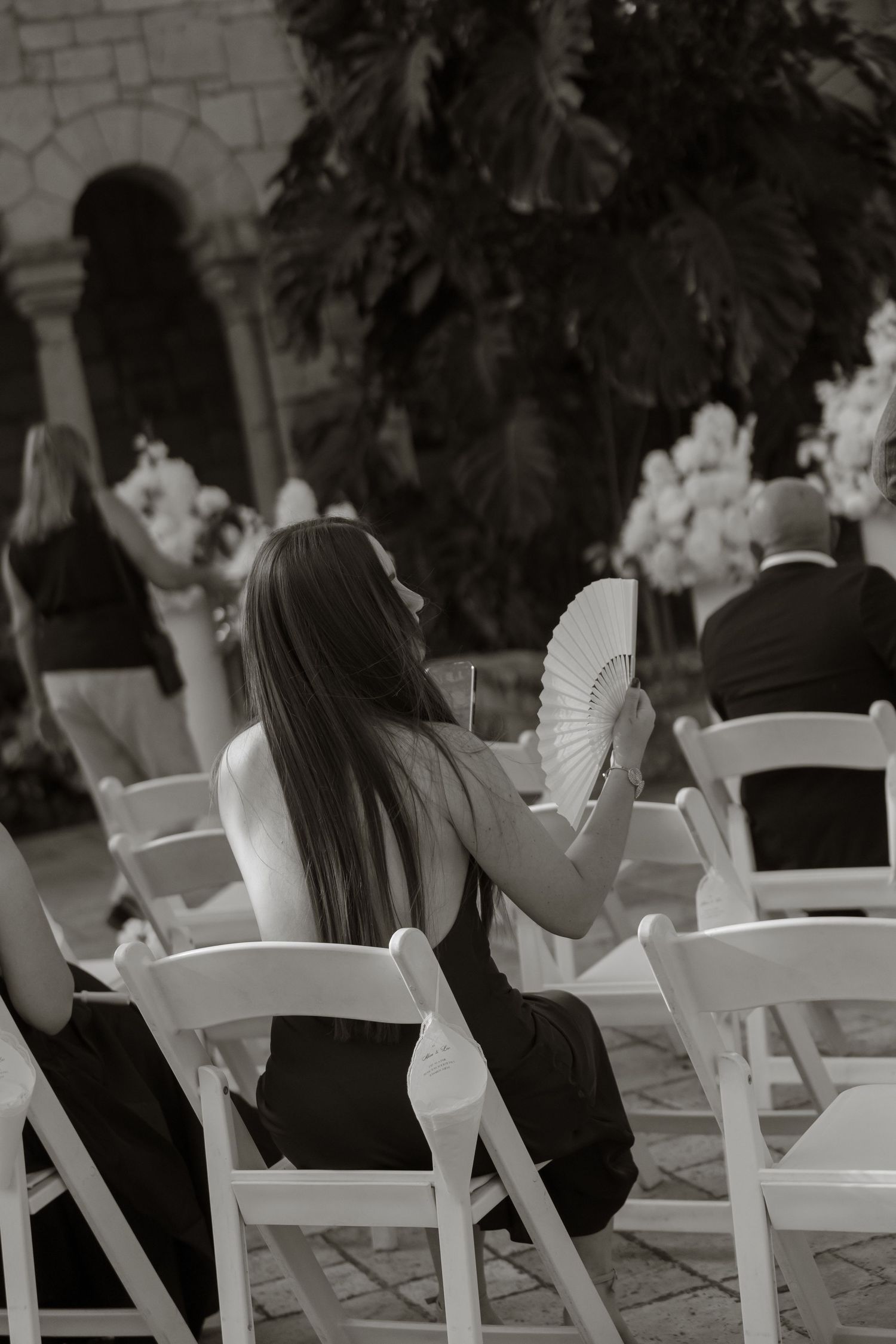 Black and white photo of a woman holding a hand fan at an outdoor wedding ceremony.
