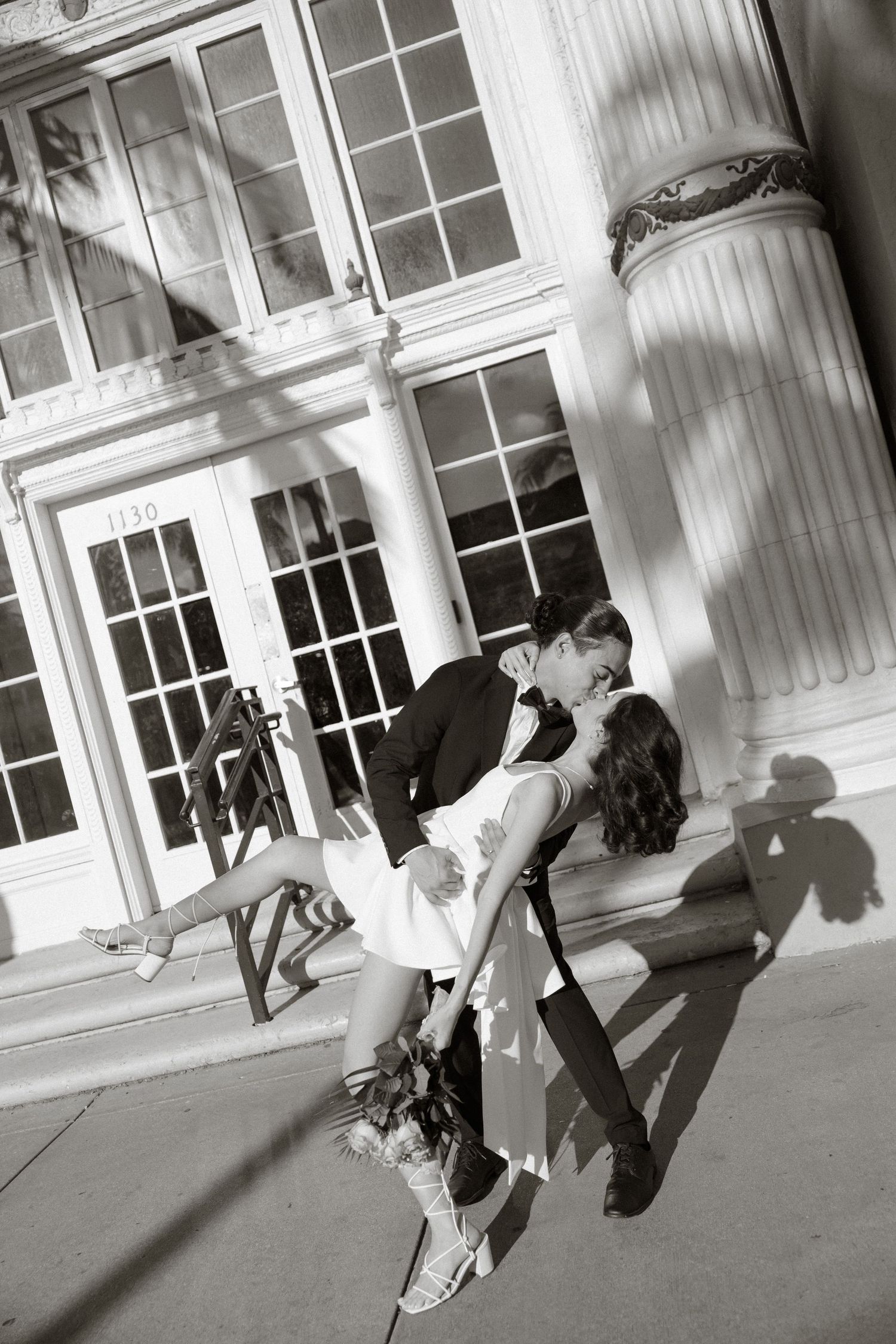 Romantic black and white wedding portrait against classic columned building with dramatic shadows.