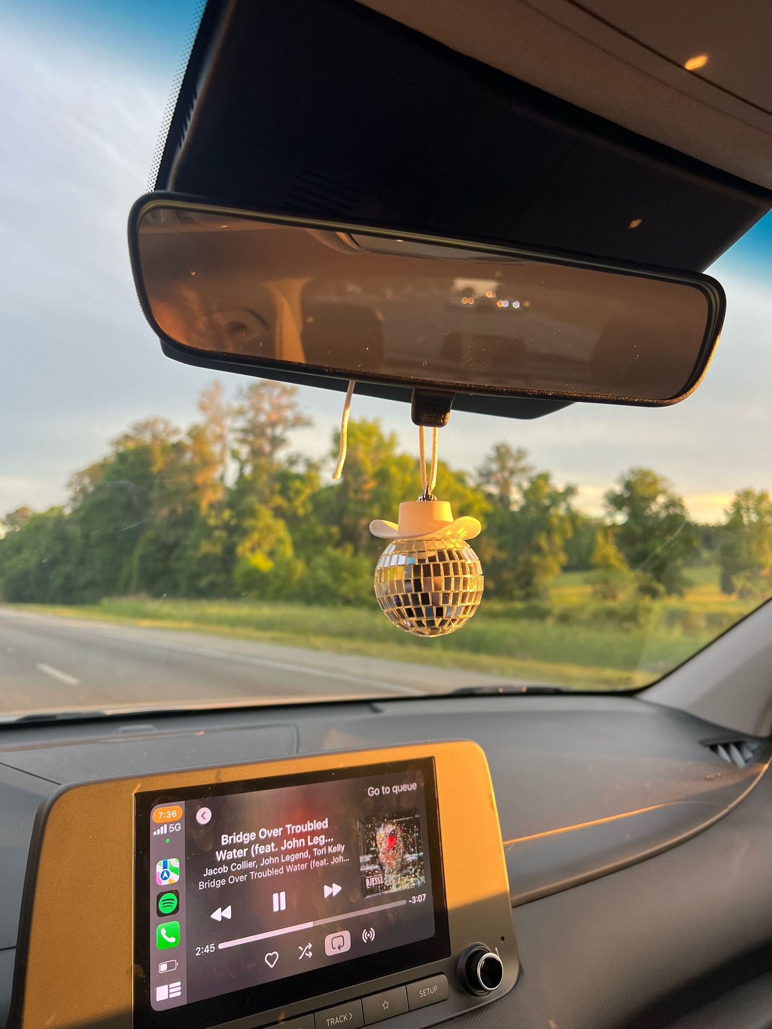 Car interior view with disco ball air freshener and infotainment display showing music player against sunset landscape.