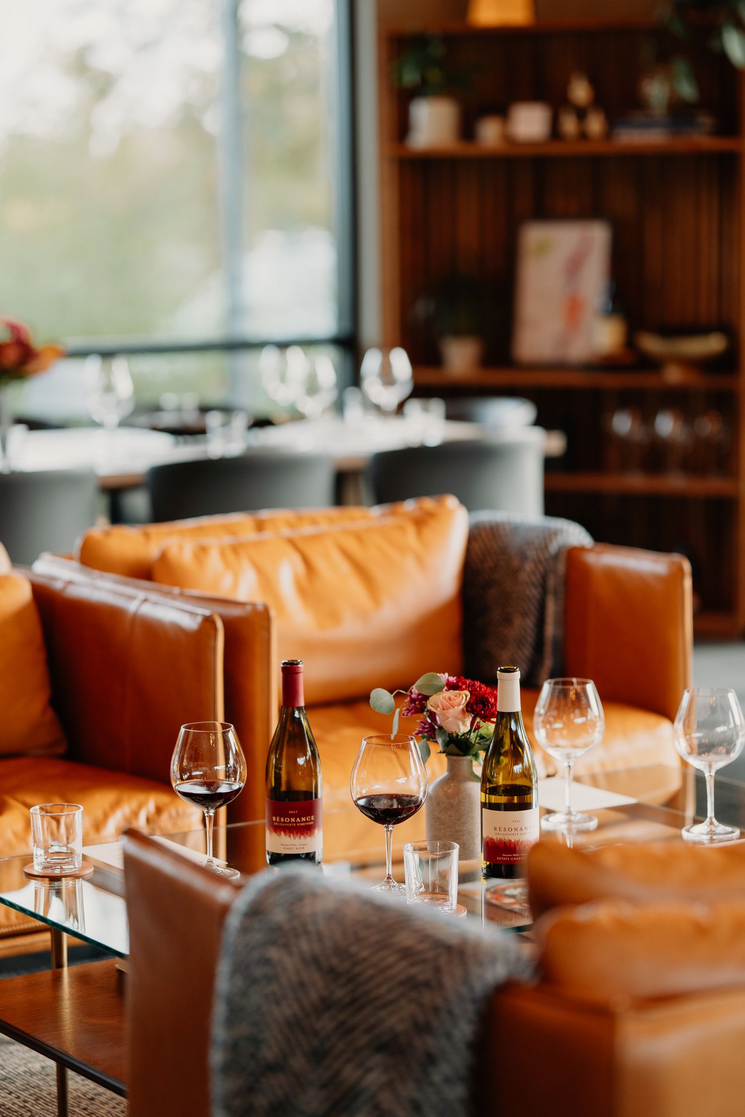 Cozy restaurant interior with leather seating, wine bottles and glasses on tables, and wooden shelving in background.