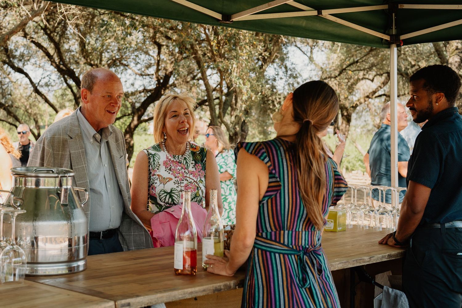 A group of people enjoying beverages and conversation at an outdoor covered venue during a sunny day.