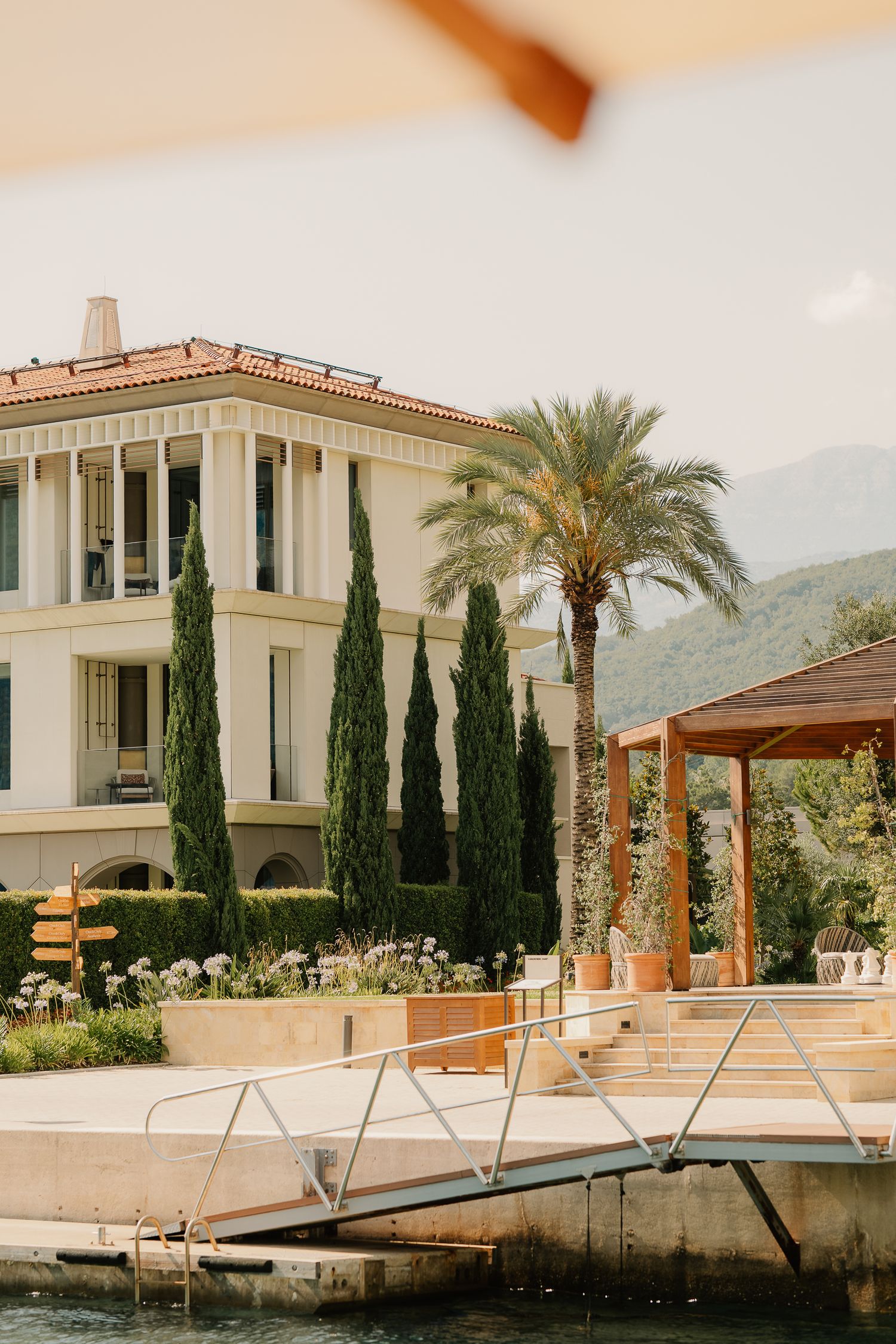 Elegant Mediterranean villa with cypress trees, palm tree, and waterfront dock on a calm lake with mountain views.