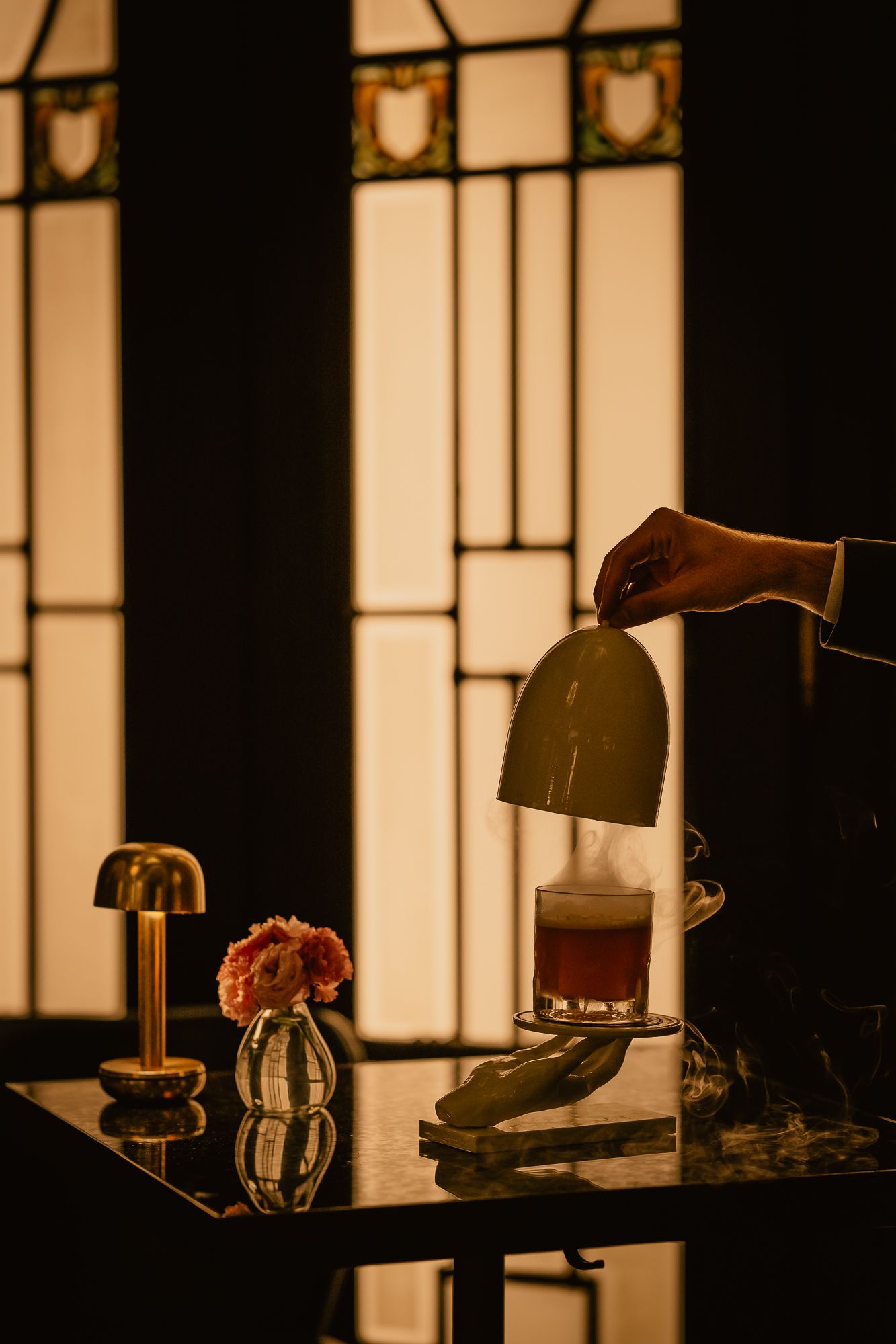 A hand lifts a dark bell cloche over a cocktail on a marble bar, beside a small lamp and pink flower vase.