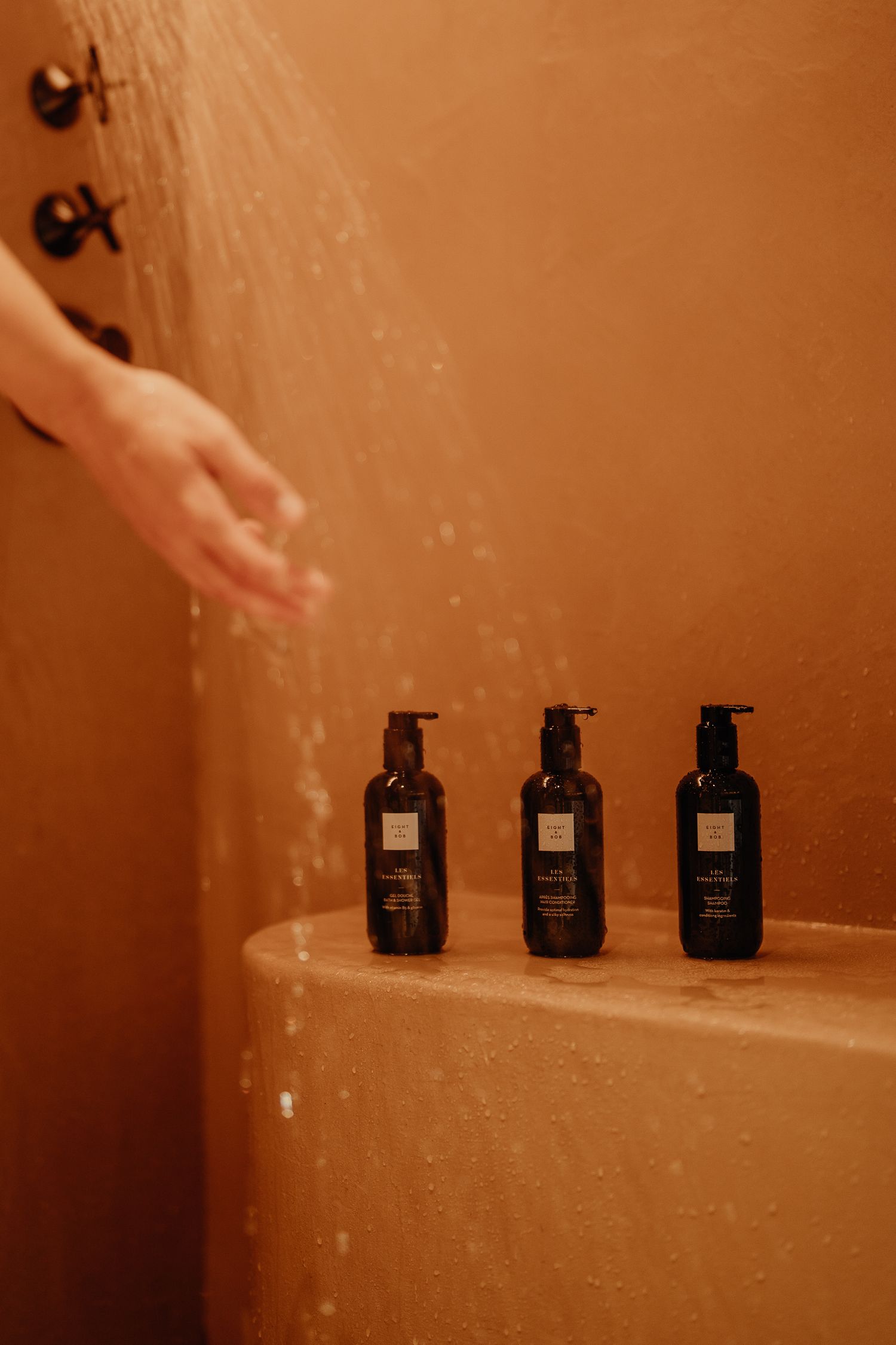 Three black pump bottles lined up on a beige shower ledge with water spray in warm lighting.