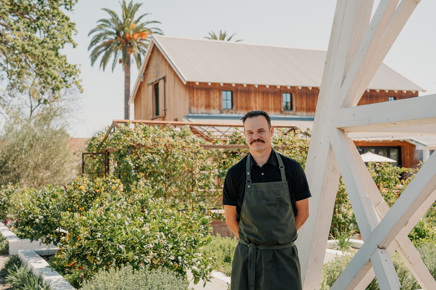 Chef in green apron stands outside a rustic farm building surrounded by lush greenery and palm trees on a sunny day.