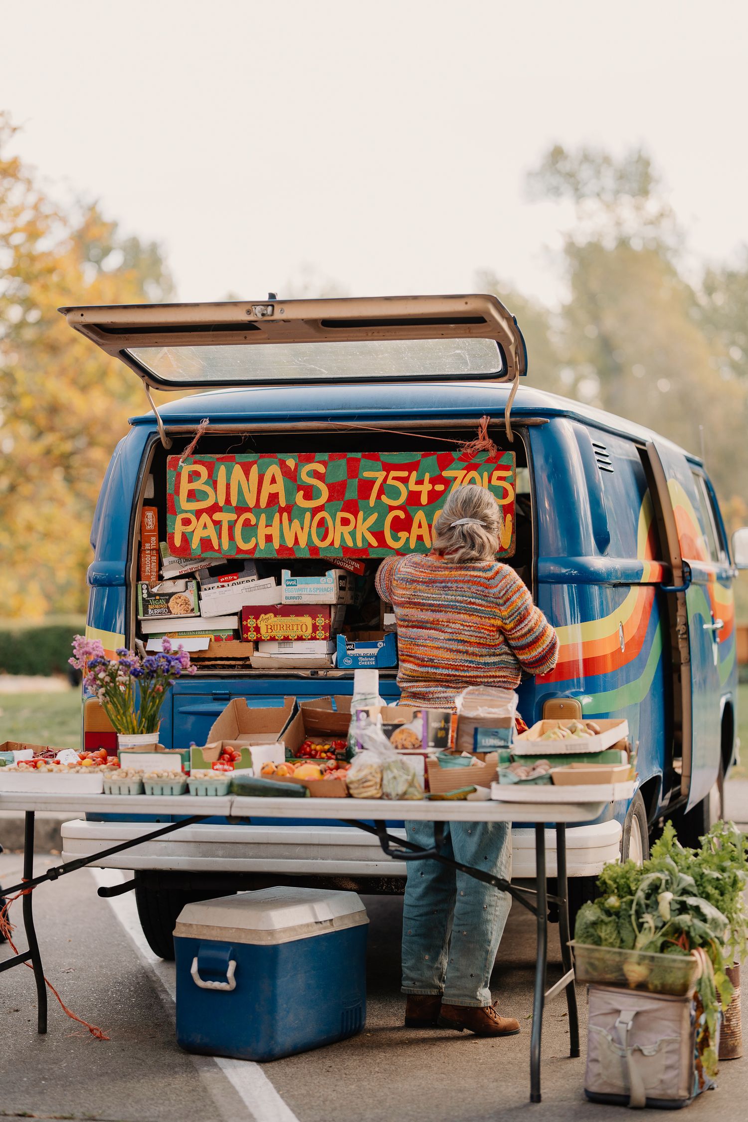 Vintage blue van with open trunk selling handmade patchwork items at an outdoor craft market with autumn trees in background.