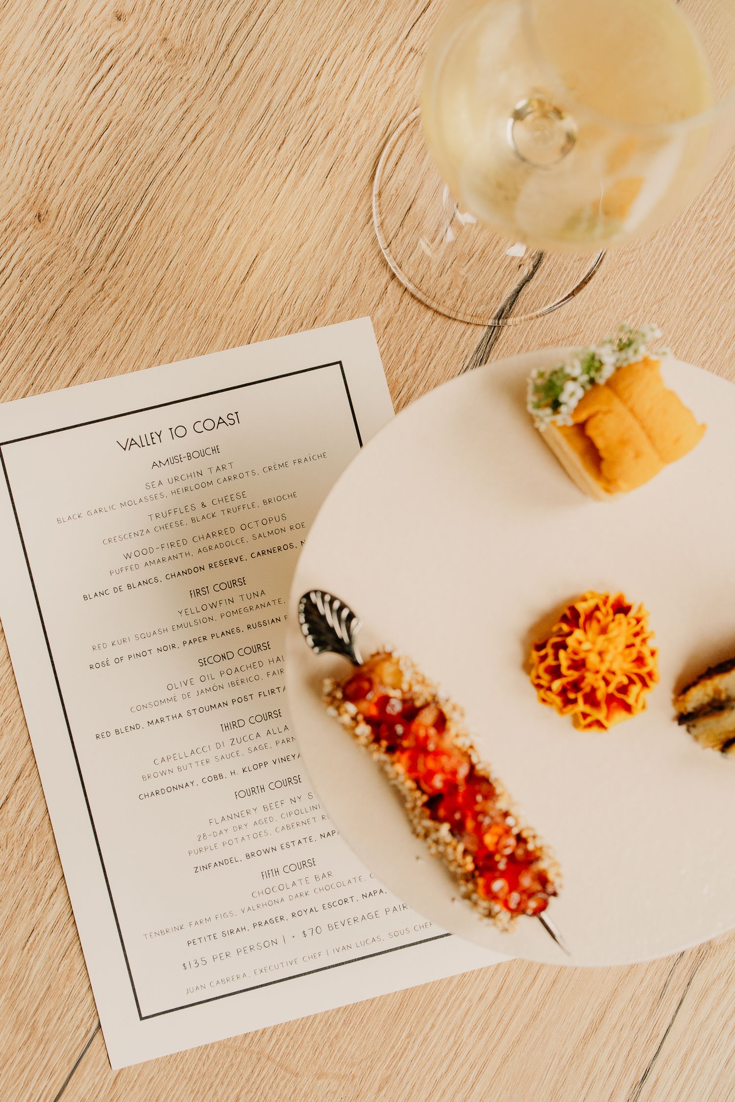A sequence of appetizer plates with orange bruschetta and menu cards on a light wooden table surface.