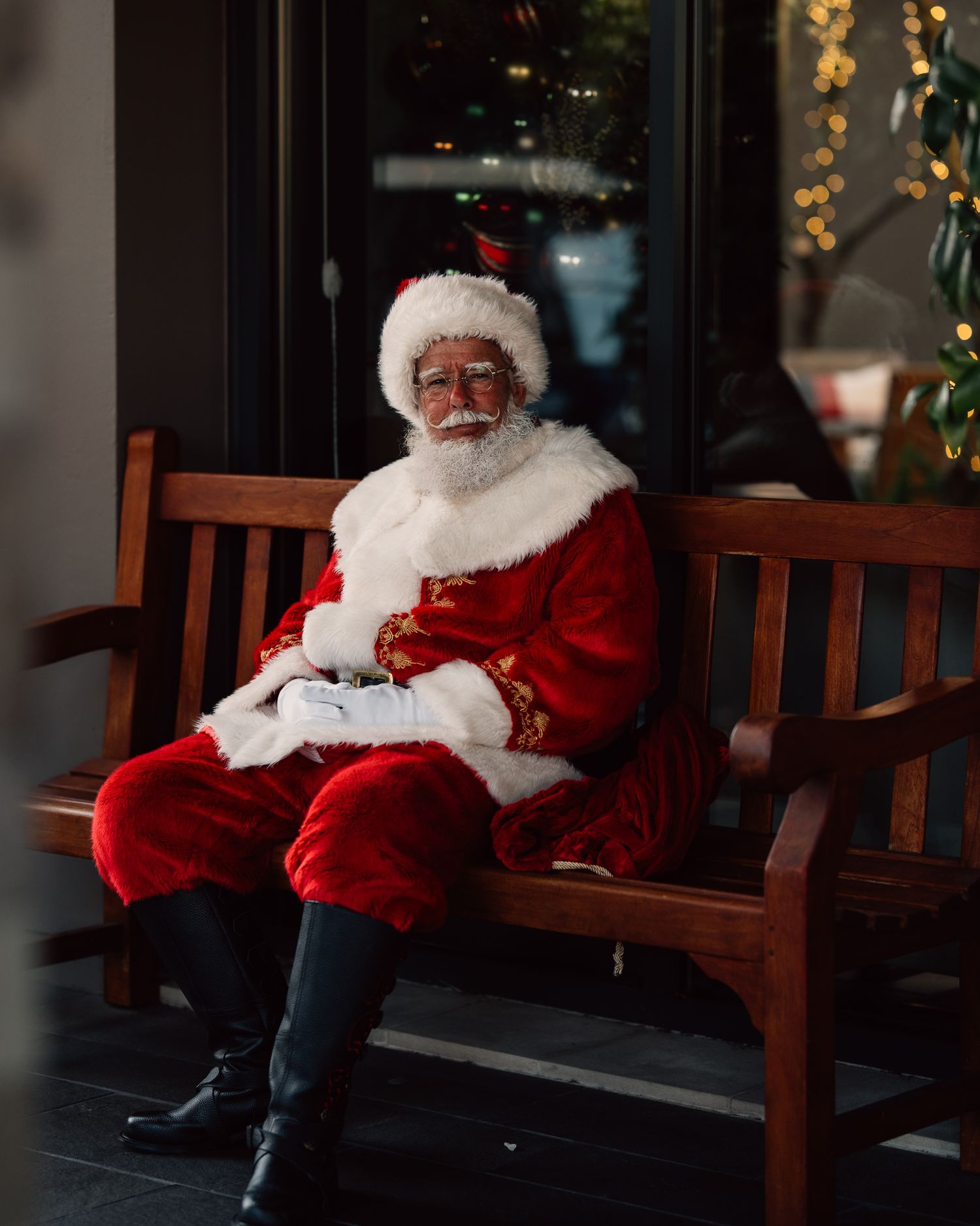 A person in a traditional Santa Claus costume sits alone on a wooden bench with Christmas decorations in the background.