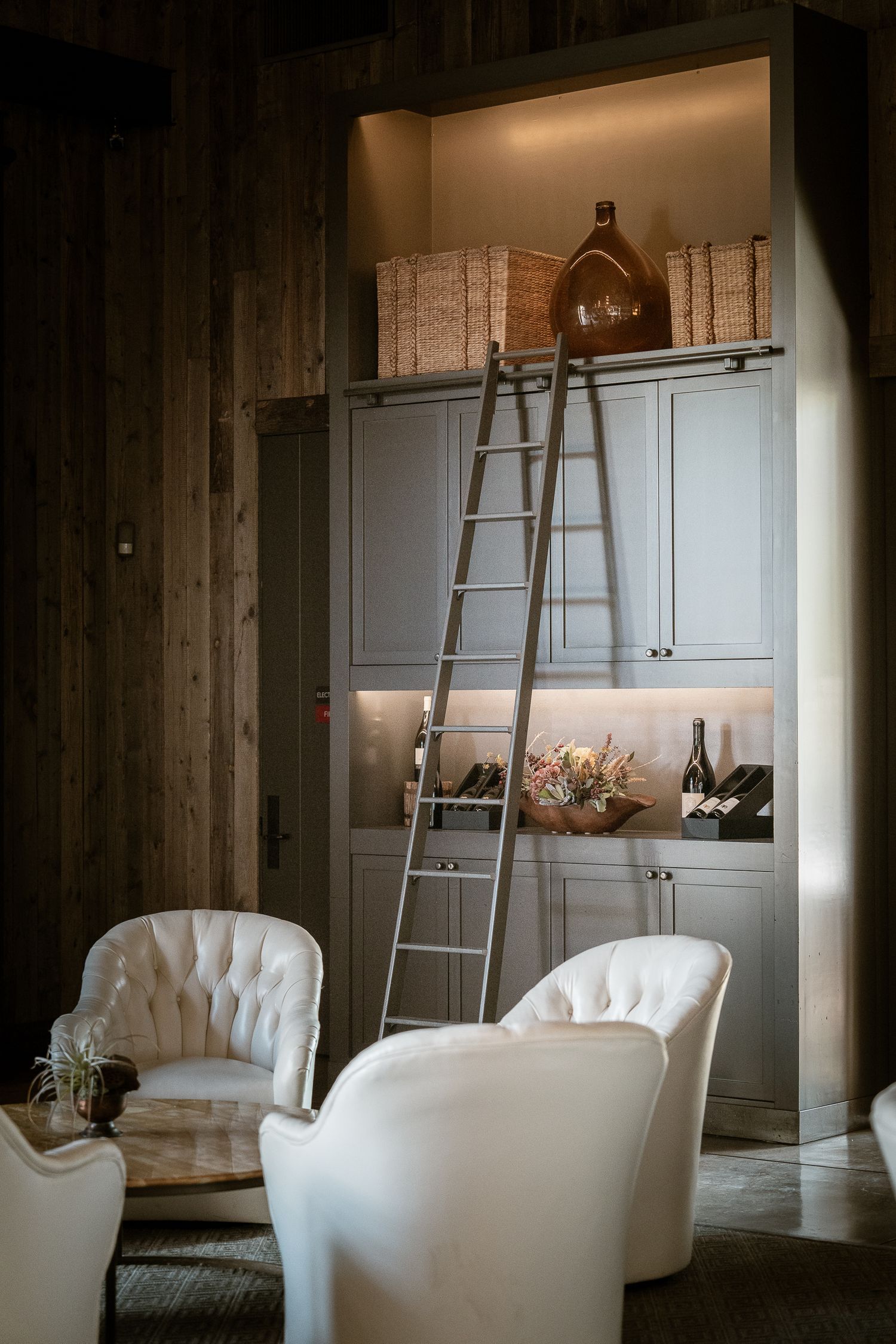 Modern living room with white tufted chairs and a tall wooden ladder leading to illuminated built-in shelving.