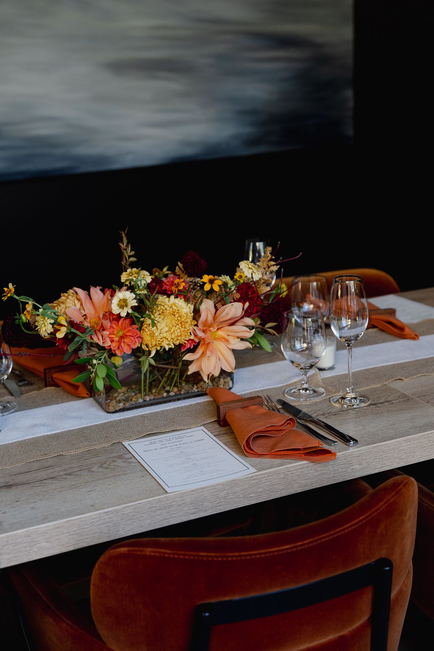 Elegant table setting with autumn floral centerpiece, wine glasses, and velvet chairs against a moody dark background.