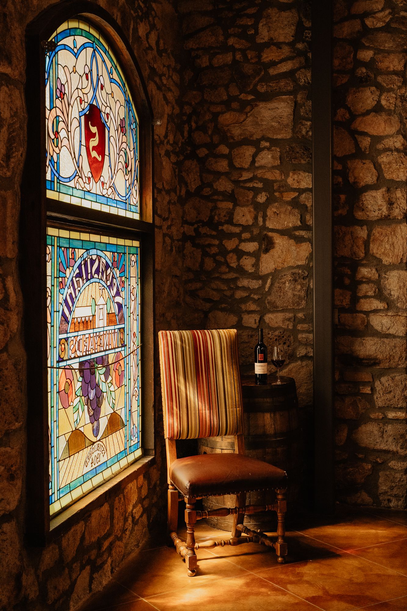 Ornate stained glass window illuminating a vintage striped chair and wine bottle in a stone wine cellar.