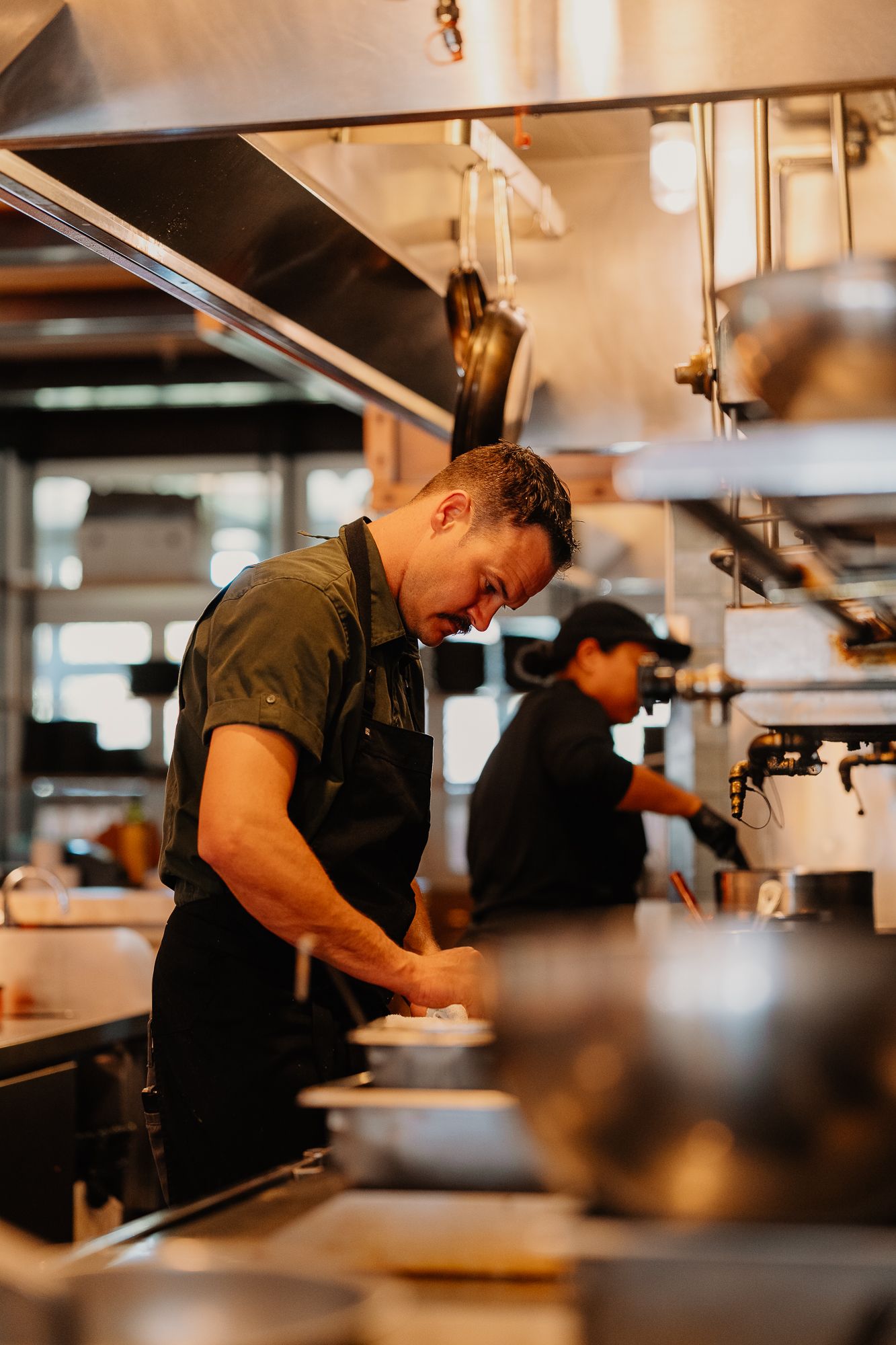 Chef in black apron smiling while plating food in a professional restaurant kitchen.