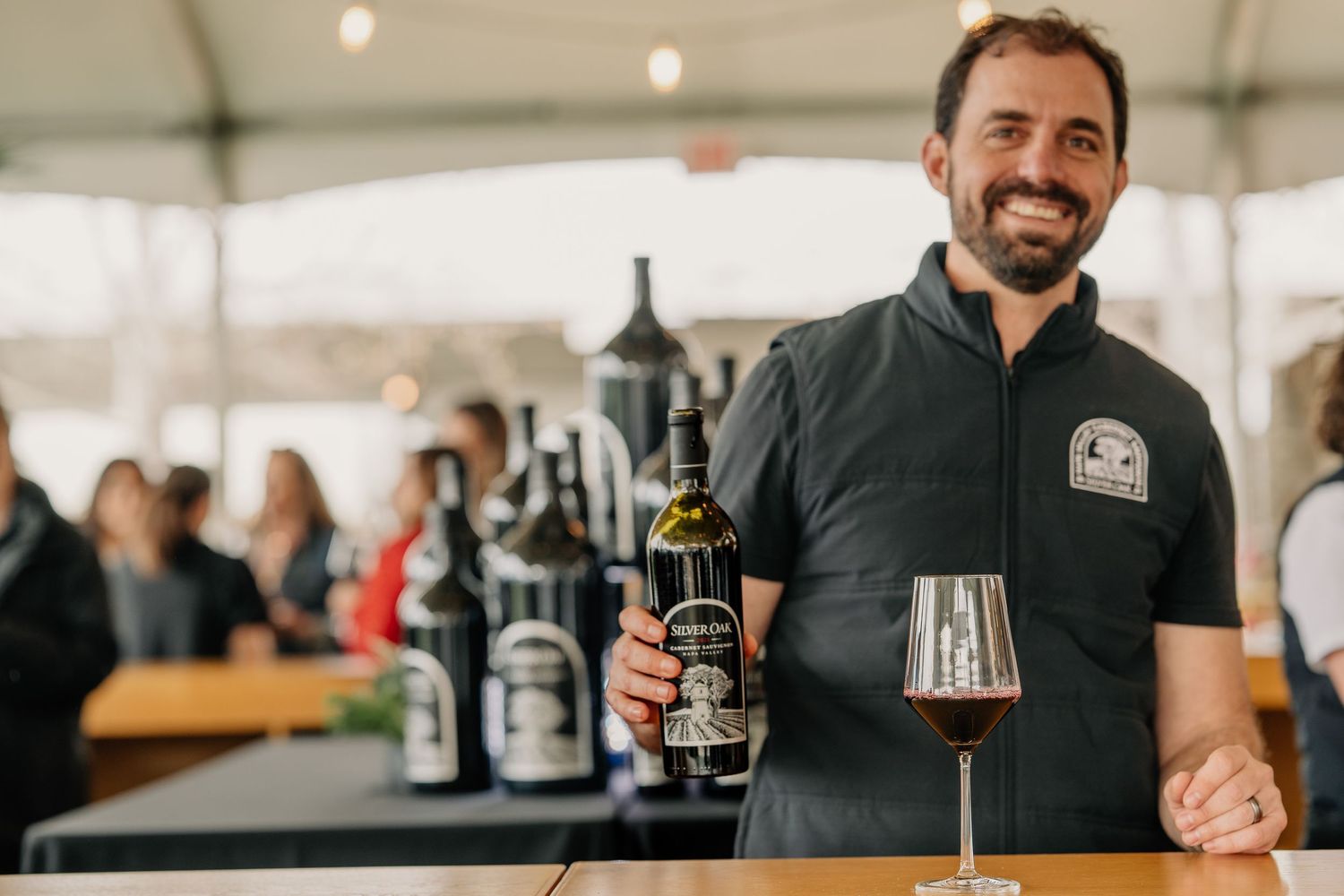 Smiling man in Silver Oak vest presents a bottle of red wine and glass at winery tasting event.