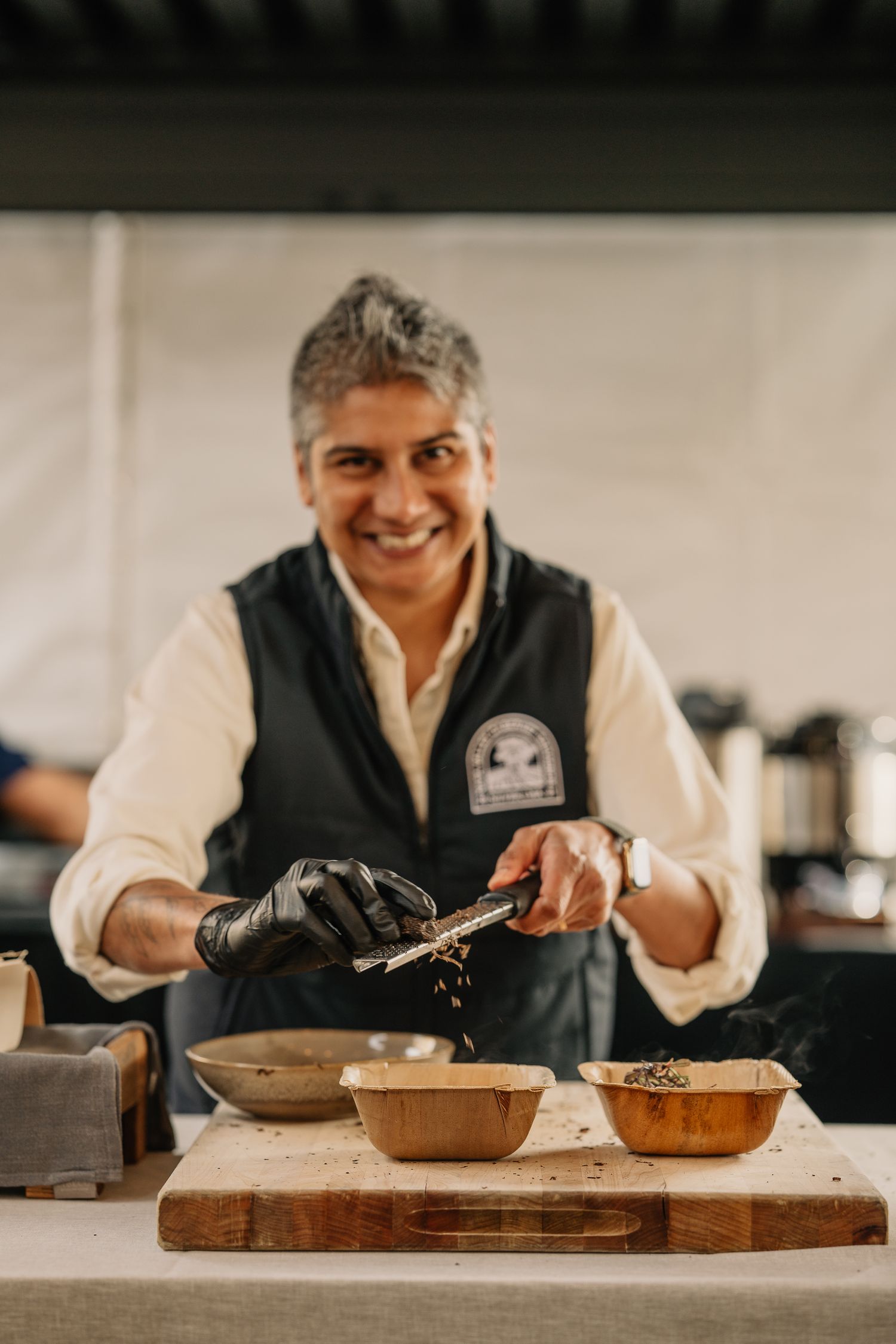 Smiling chef in black vest and gloves uses tongs to plate food into wooden bowls at an outdoor culinary event.