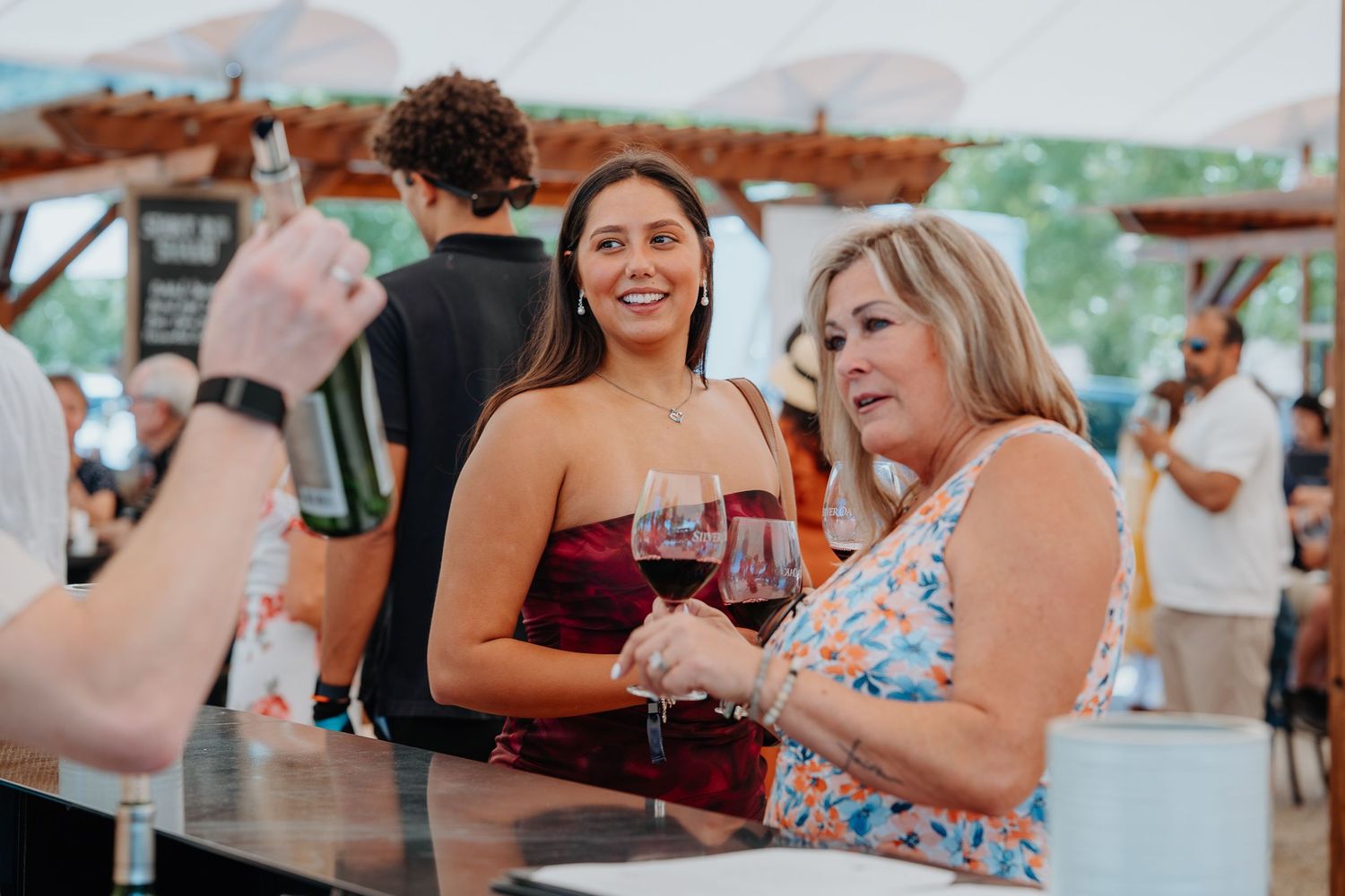 Two women smiling and holding red wine glasses at an outdoor wine tasting event under a white tent.