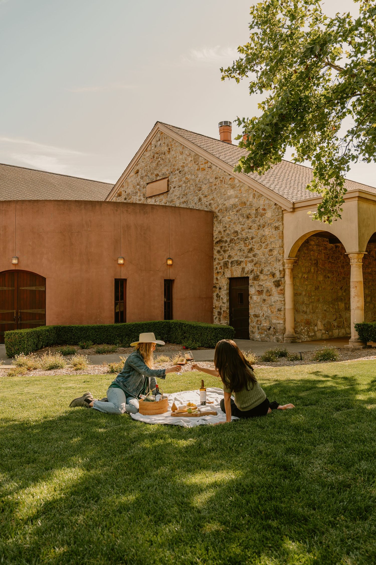 Two women enjoying a picnic with wine on green lawn outside a rustic stone winery building.