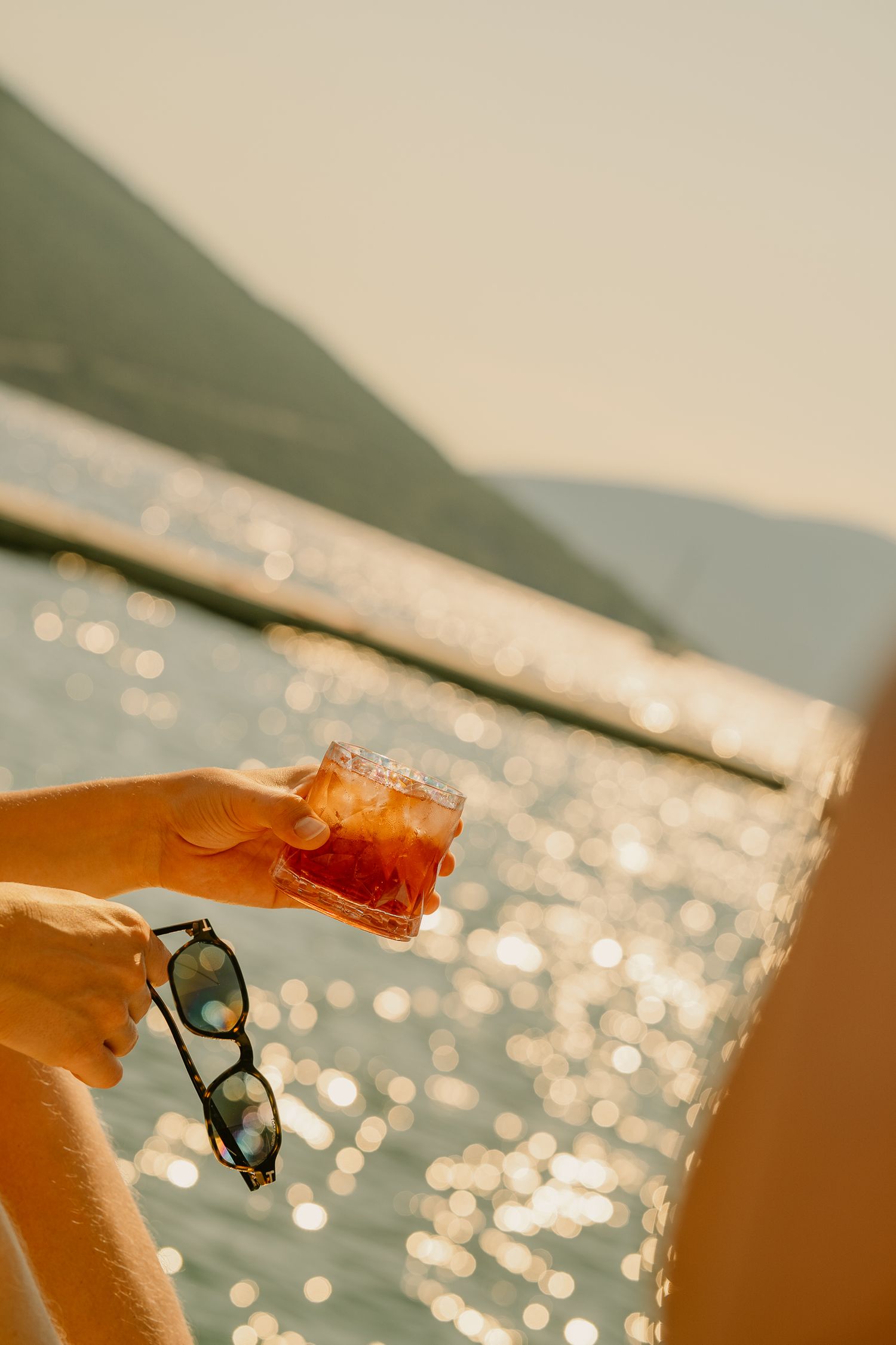 Person holding a cocktail with ice and sunglasses by a sparkling sunlit sea with mountain backdrop.