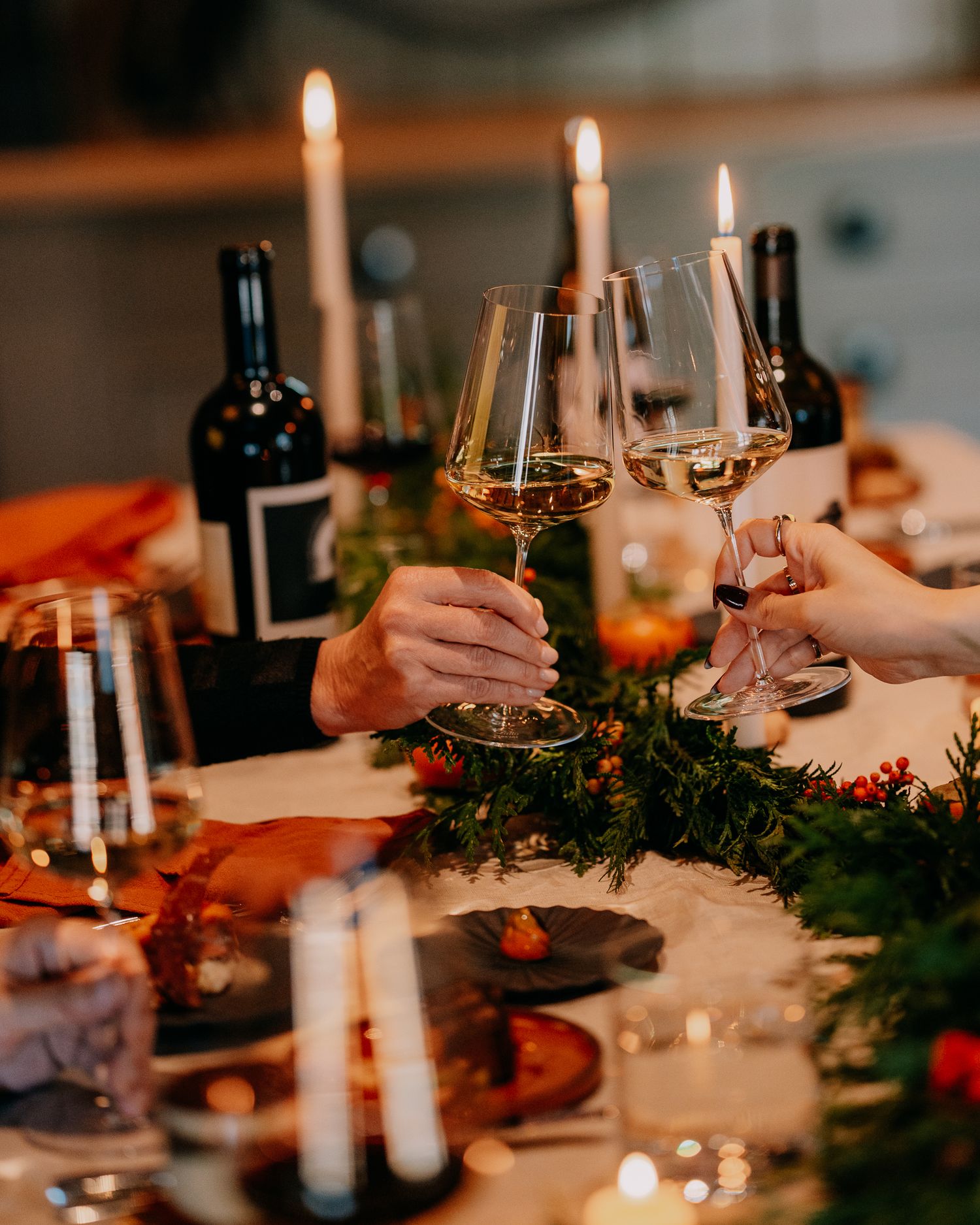 Festive holiday dinner table setting with lit candles, wine glasses, and evergreen garland decorations in warm lighting.