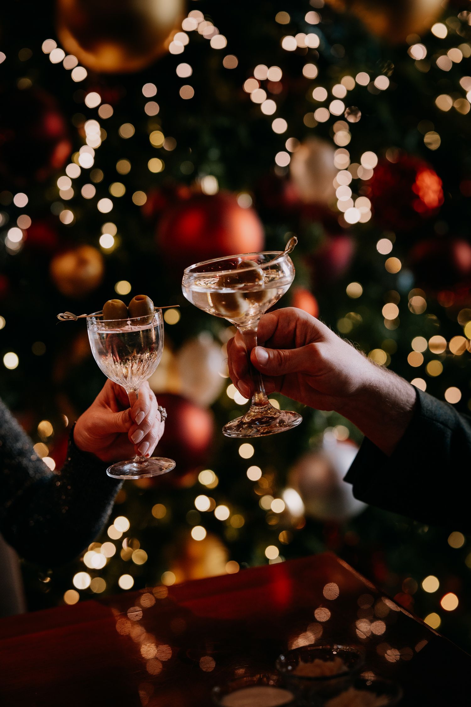 Hands holding vintage champagne coupes against blurred Christmas lights and bokeh background creating a festive atmosphere.